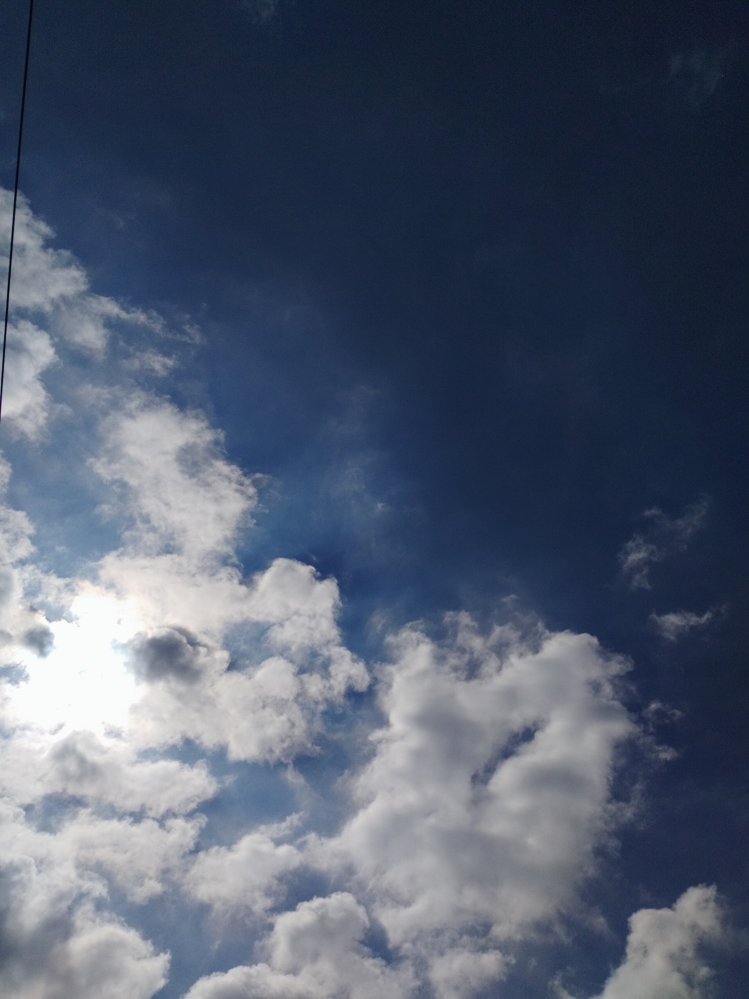 A dark blue sky is partially obscured by bright white clouds. The sun is visible through a gap in the clouds on the left side of the image, casting a bright light. A thin black line, likely a utility wire, runs vertically down the left edge of the frame. The image appears to have been taken from a low angle, looking upwards. No specific landmarks in Lhokseumawe, Indonesia are visible.