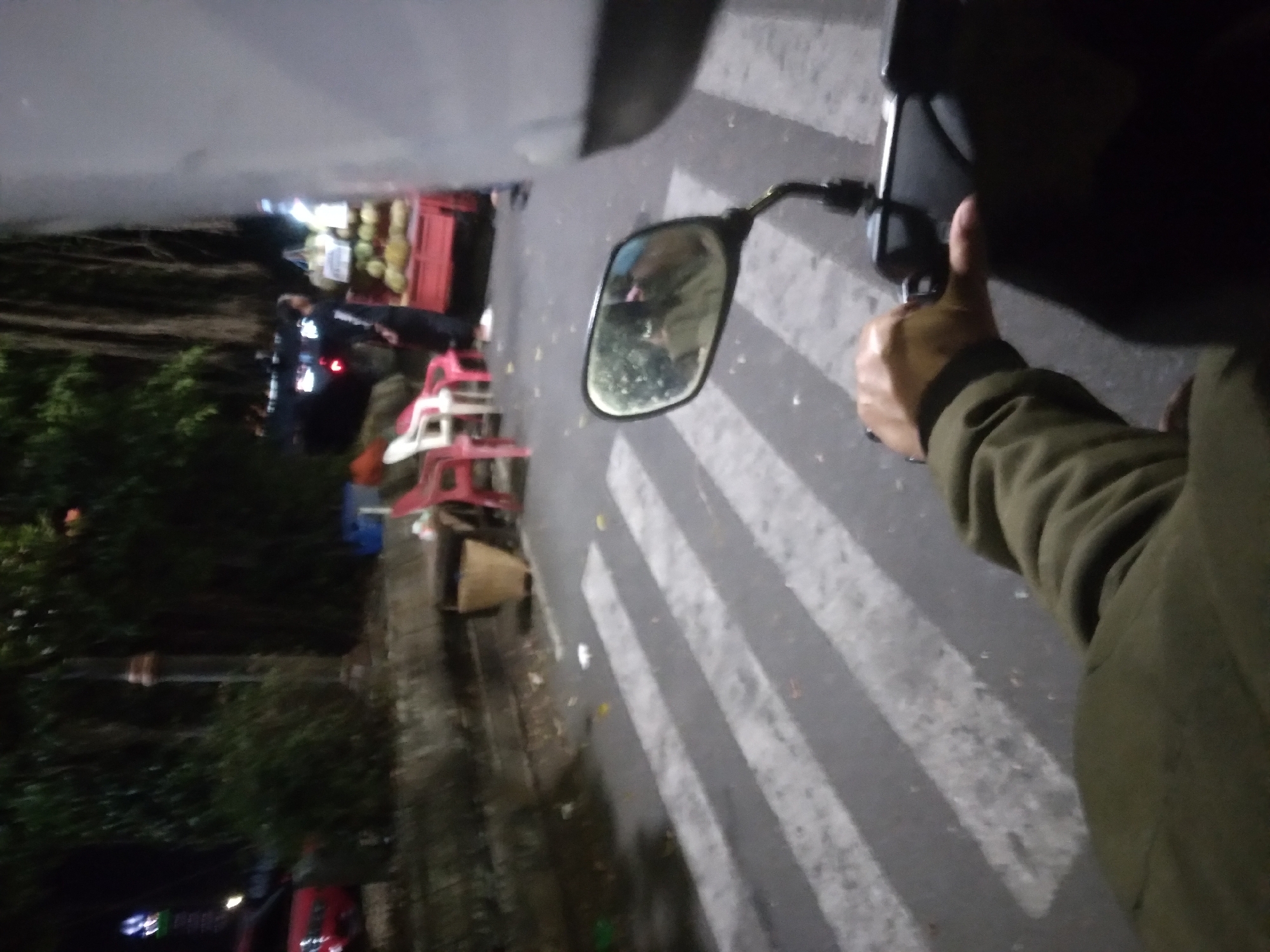 The image captures a street scene at night in Purwokerto, Indonesia, likely near a busy area given the presence of a street vendor and pedestrian crossing. In the foreground, a hand on a motorcycle handlebar is visible, suggesting the photo was taken from a moving vehicle. A side mirror reflects a blurry image of the rider and their surroundings.
Further back, a pedestrian crossing with white stripes is visible on the asphalt road. To the left of the road, a wall is covered in greenery, and a street lamp stands tall. Beyond the wall, a red car is partially visible.
On the right side of the road, a fruit vendor's cart is illuminated, displaying a variety of fruits, possibly durians. A person in dark clothing, presumably the vendor, stands behind the cart. Several plastic chairs, one red and one white, are placed near the vendor's cart.
The overall scene depicts a typical Indonesian street at night, with a mix of urban elements and the hustle of street commerce.