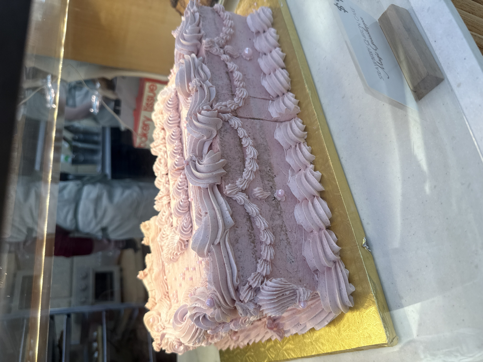 A large, rectangular cake with light pink frosting is prominently displayed within a glass case at a bakery in Saint Petersburg, United States of America. The cake, situated on a textured gold foil cardboard cake board, is heavily adorned with piped light pink frosting forming intricate swirls, ruffles, and garland-like patterns, further embellished with small, spherical pink sprinkles. A clear tent-style sign with a wooden base in the foreground identifies the item as "STRAWBERRY CHIFFON CAKE" and also mentions "Magic Grasshopper," with a partial price indication of "$$." The glass display case offers a reflected and partially visible view of the bakery's interior beyond. Two individuals are discernible in the background through the glass: one person, wearing a white apron over a red shirt, is partially visible and facing away in what appears to be a kitchen area with metal shelving and an oven. Another less distinct figure in a light-colored uniform is also present. A red and white box marked "bakery tissue" is visible in the background, along with various kitchen items. The counter surface beneath the display case is a speckled white.