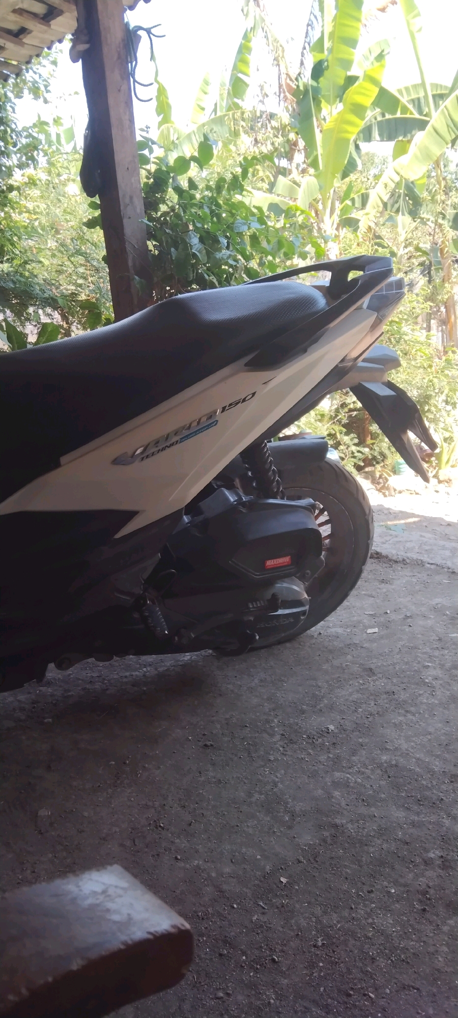 The image shows a side view of a white and black motorcycle parked outdoors. The motorcycle is the primary subject, with its seat, rear fender, and part of the engine casing visible. The bike appears to be parked on a dirt or gravel surface, with a wooden structure and lush green foliage in the background. The environment suggests a tropical or subtropical setting, possibly Wadang, Indonesia, given the presence of banana trees and dense vegetation. The lighting indicates it is daytime, with sunlight filtering through the leaves. There are no people or animals in the frame. The overall scene is static and appears to be an everyday outdoor setting.