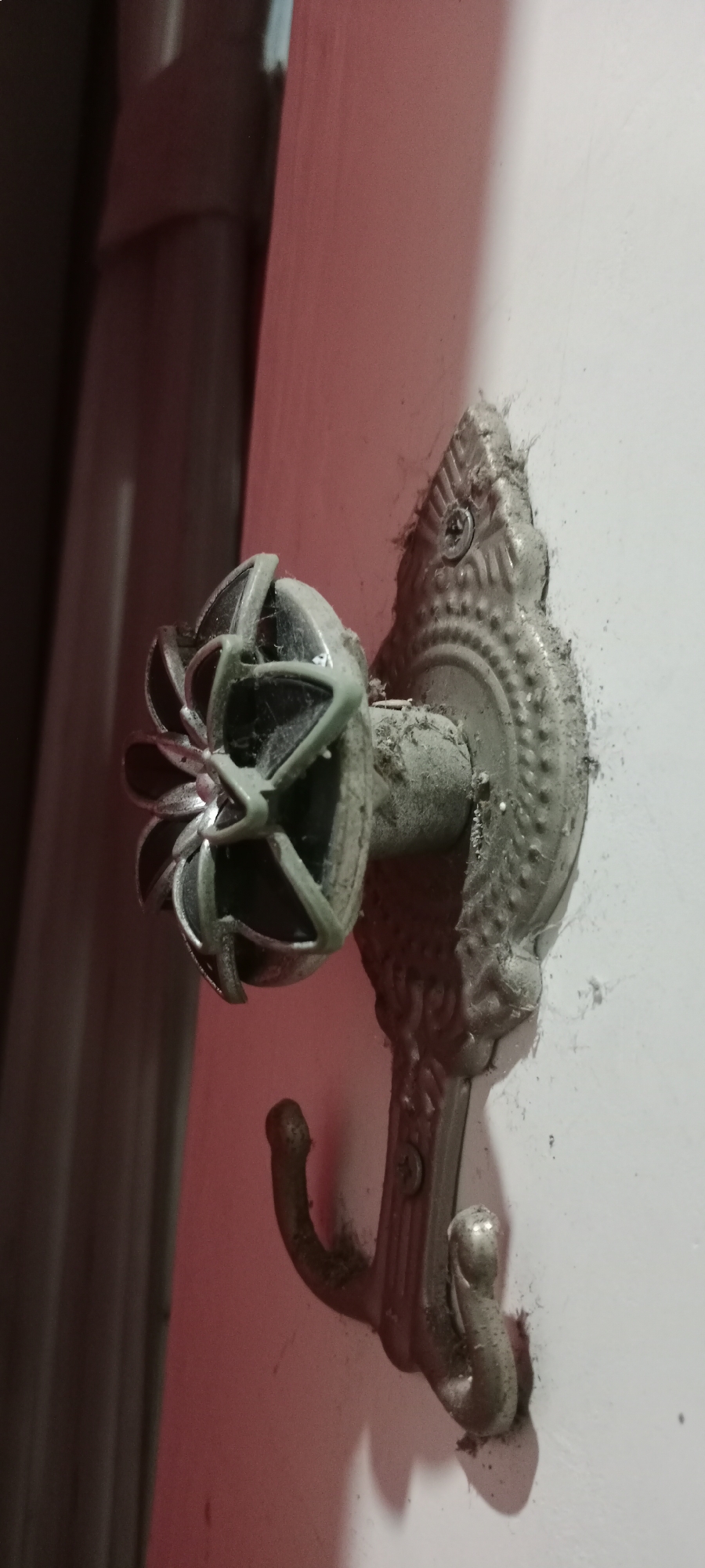 The image shows a close-up of a silver-colored metal hook mounted on a white wall. The hook is ornate, with a decorative backing that has a patterned texture. Attached to the hook is a green decorative element with a floral or starburst shape, which appears to be made of plastic or a similar material. The entire object, including the hook and the decorative piece, is covered in a layer of dust and cobwebs, indicating it is likely old or has not been cleaned for some time. The lighting in the image is dim, with a reddish hue cast on the left side, possibly from an external light source or reflections. The background on the left is dark and appears to be a vertical surface, possibly a curtain or a door. The image is a still shot and does not depict any ongoing action or interaction. There is no identifiable location in the city of Soko, Indonesia, visible in the image.