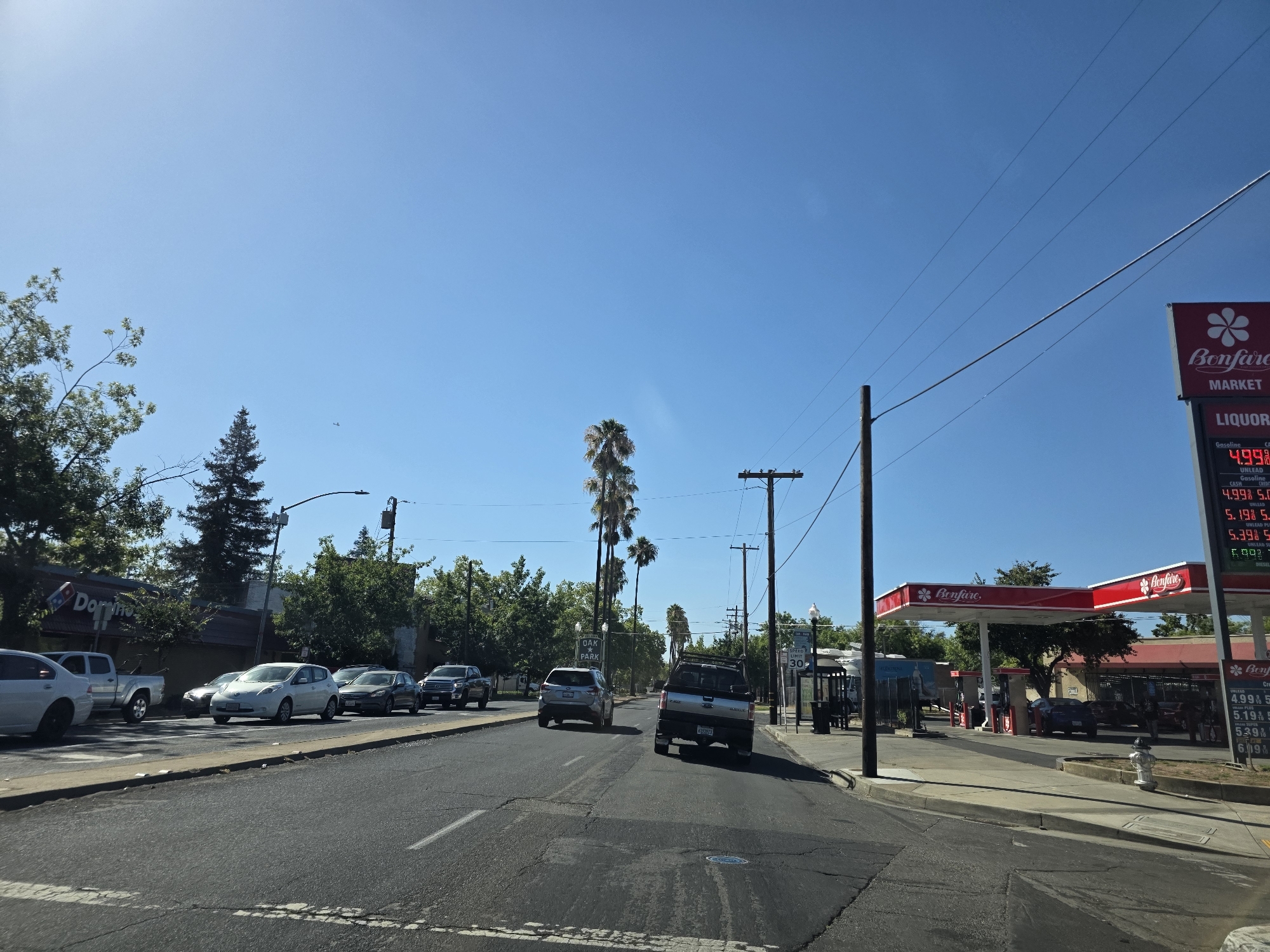 This is a horizontal, eye-level, medium shot taken on a clear, sunny day. The scene depicts a street in Sacramento, California, United States of America, with traffic and a gas station.
In the foreground, the asphalt road shows some wear and tear with visible cracks and white lane markings. To the left, a concrete sidewalk is visible, followed by parked cars and trees. A silver sedan is parked on the left side of the road. Further down the street, several cars are driving in the same direction as the camera, including a silver Nissan Leaf, a dark grey sedan, and a black pickup truck. A silver pickup truck is also visible further down on the left.
On the right side of the street, a Bonfare market and gas station is prominent. A red and white sign displays gas prices, with unleaded gasoline priced at $4.99 and premium at $5.39. Several cars are at the gas pumps. A bus stop shelter with a bench is also on the right side of the road, along with a speed limit sign indicating 30 mph.
In the background, trees and buildings line the street. A Domino's Pizza sign is visible on the left. Palm trees are scattered throughout the scene, including a cluster of tall palm trees in the middle distance. The sky is clear blue, with some power lines crisscrossing the upper portion of the image. A small airplane is visible in the sky on the left side.