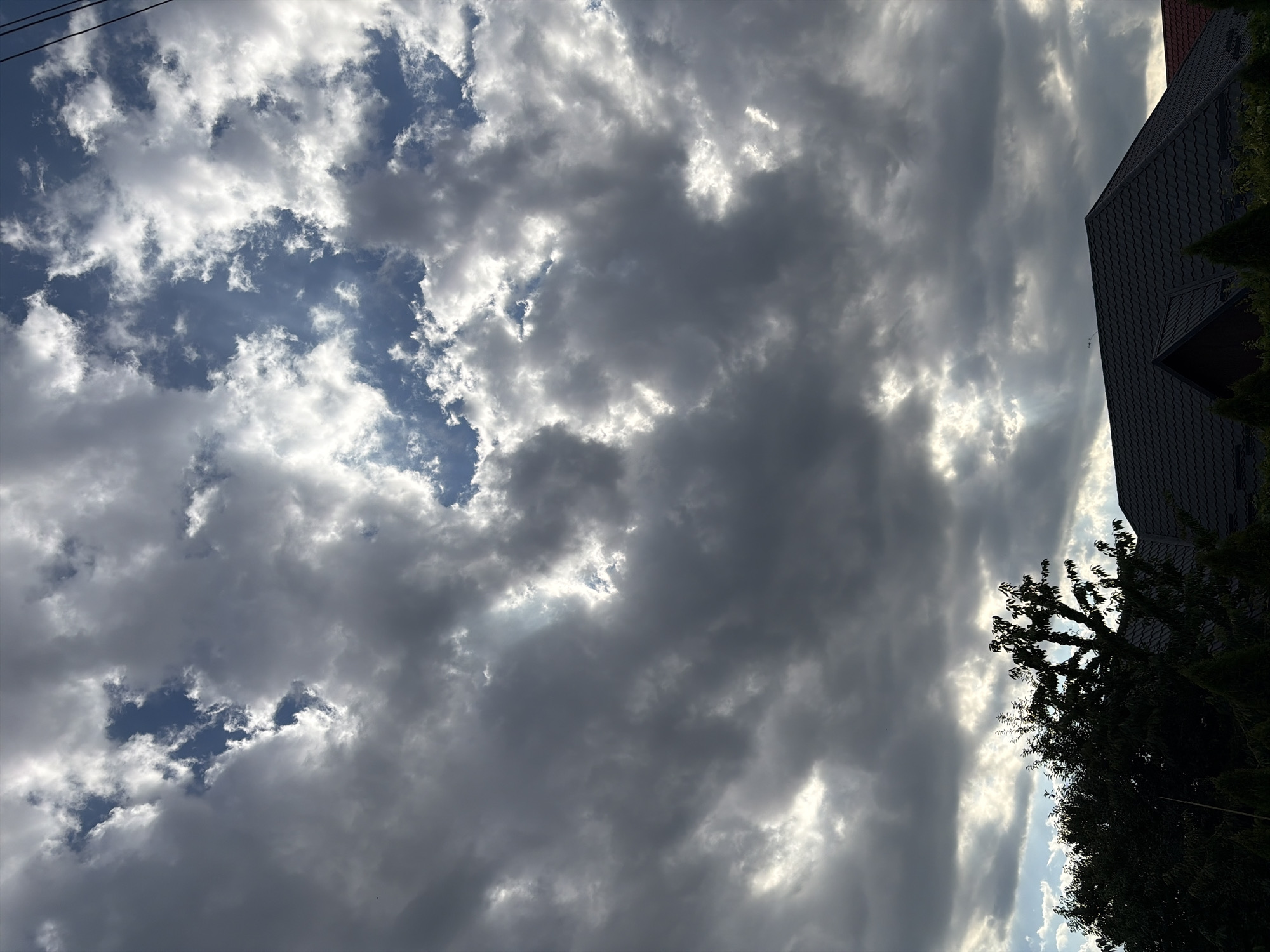 This is a low-angle, portrait-oriented photograph taken outdoors in Oxentea, Moldova. The image captures a dramatic sky filled with a mix of dark, heavy storm clouds and bright, sunlit patches. The sun's rays are piercing through the clouds, creating a dynamic interplay of light and shadow.
In the foreground, the dark, textured roof of a building, likely a house, is visible on the right side of the frame, extending diagonally from the bottom right corner upwards. Adjacent to the roof, a dense, dark green tree or shrubbery is silhouetted against the bright sky.
The background is dominated by the sky, which is the main subject. The clouds are varied in texture and density, with some appearing almost solid and dark, while others are wispy and illuminated. The patches of blue sky visible between the clouds suggest that the storm is either approaching or receding.
The lighting suggests it is either late afternoon or early morning, as the sun is low enough to create dramatic rays. The weather appears to be mixed, with the potential for rain indicated by the dark clouds, but also sunshine breaking through. There is no visible text in the image. The overall mood of the image is one of powerful natural forces at play, with a sense of foreboding mixed with hope from the sunlight.