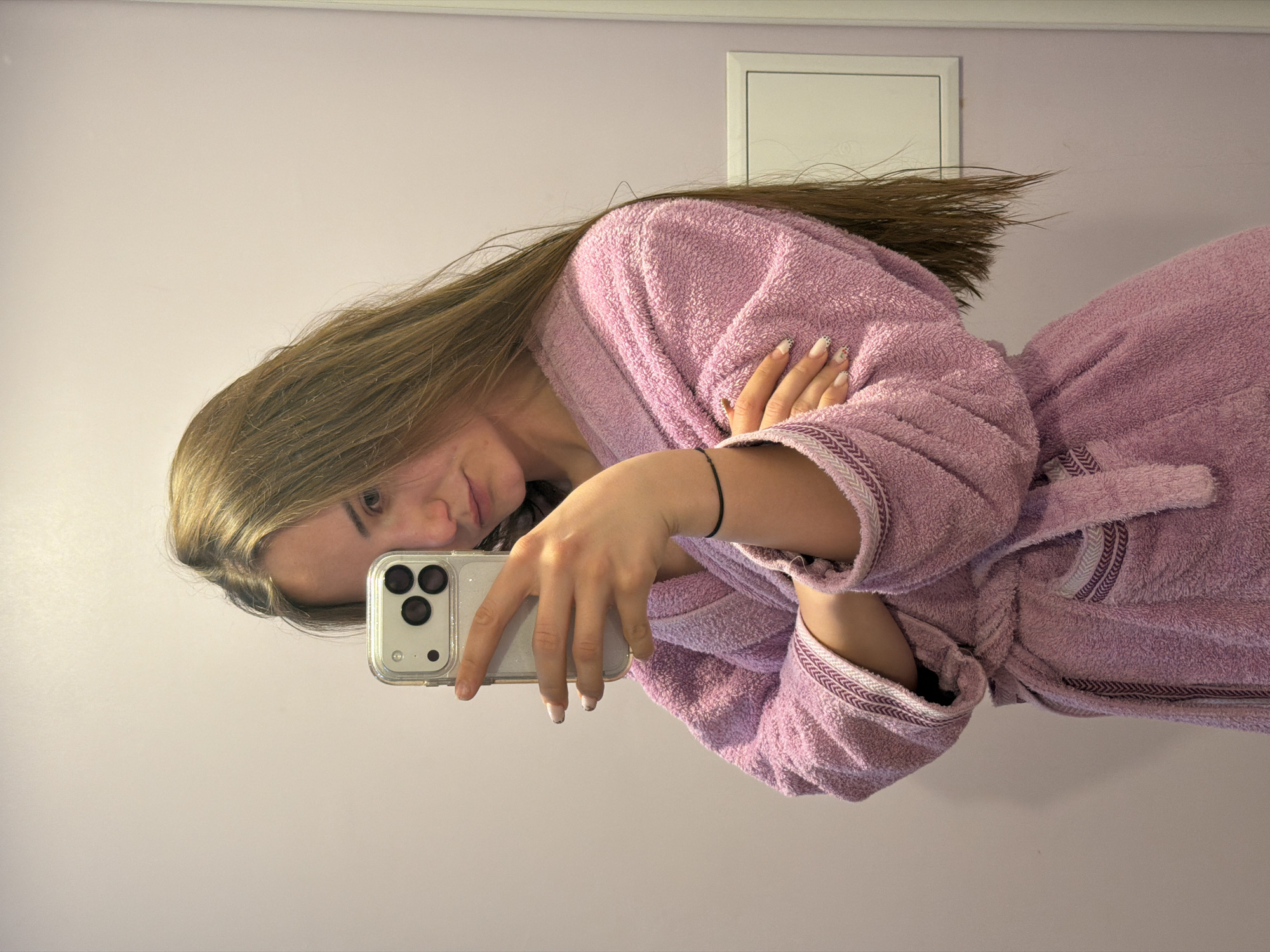 An indoor mirror selfie captures a young woman lying on her side, appearing relaxed in what seems to be a personal space within Buhovo, Bulgaria. She has long, straight light brown hair and is wearing a soft, purple terry cloth bathrobe with textured patterned trim on the cuffs, collar, and tie. Her nails are manicured, featuring a subtle white base with black speckles or patterns on some fingers.
She is holding a light-colored smartphone with a clear case and a distinct triple camera setup, using it to take the photograph reflected in the mirror. Her left arm, adorned with a thin black string bracelet, is bent, supporting her head, while her right arm is crossed over her chest. She looks towards the phone's lens with a neutral to slightly smiling expression.
The background consists of a plain, light-colored wall, possibly pale lavender or off-white. A white rectangular vent or access panel is visible on the wall above her body. The lighting is soft and even, suggesting an indoor setting without direct sunlight. There is no visible text in the image. The overall impression is one of casualness and comfort.