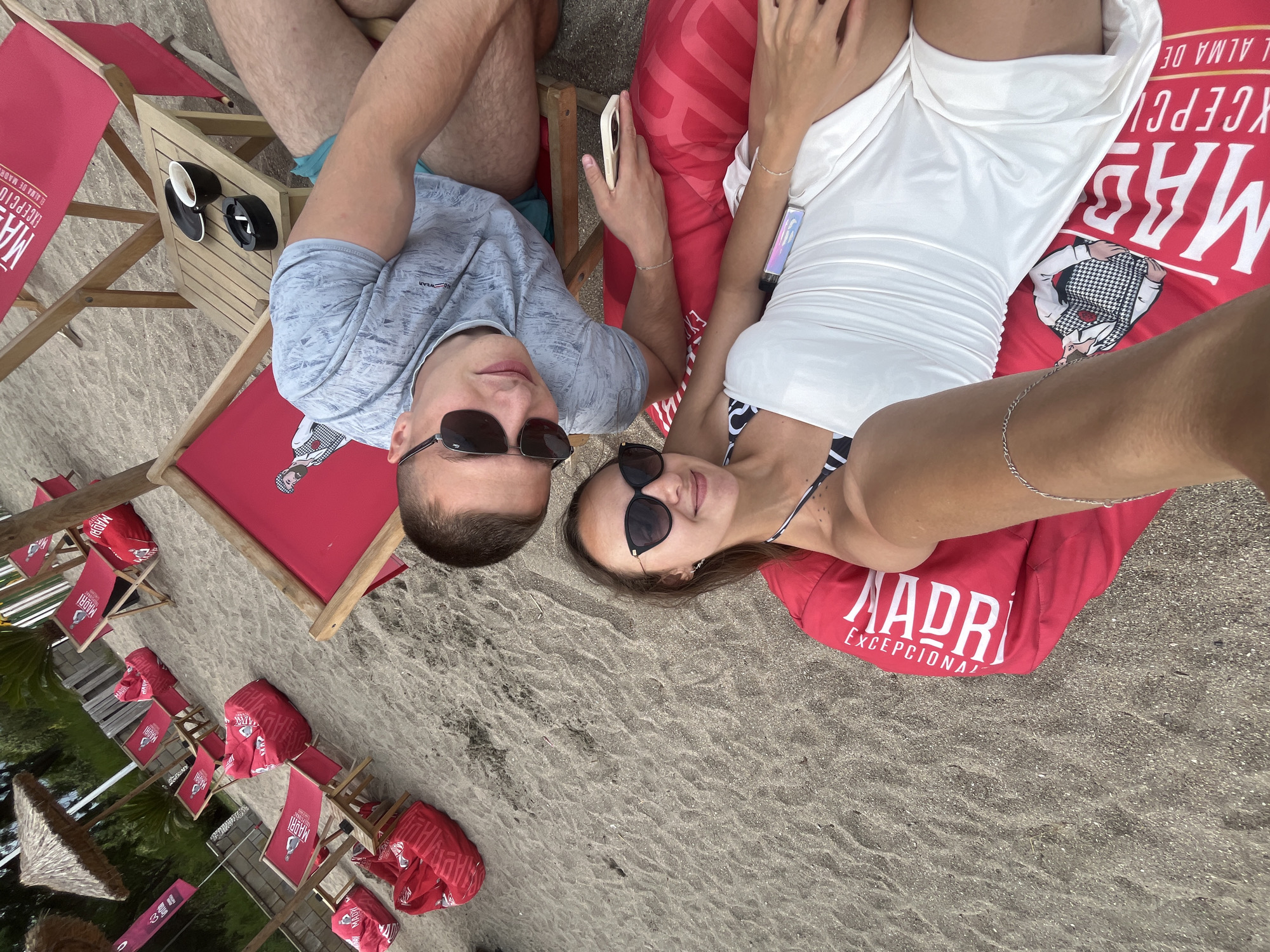 In Burgas, Bulgaria, a man and woman are captured in a selfie on a sandy beach. The couple is positioned on large, red beanbag chairs, one of which is branded with the text "MADRI EXCEPCIONAL". The man, wearing a grey t-shirt and blue shorts, is lying down, looking at the camera, with sunglasses on. The woman, in a white dress and a bikini top, is also lying down, facing the camera with sunglasses. Behind them, several red beach chairs and umbrellas are visible, suggesting a beach resort or club. The ground is covered in sand, and the overall atmosphere is relaxed and sunny.