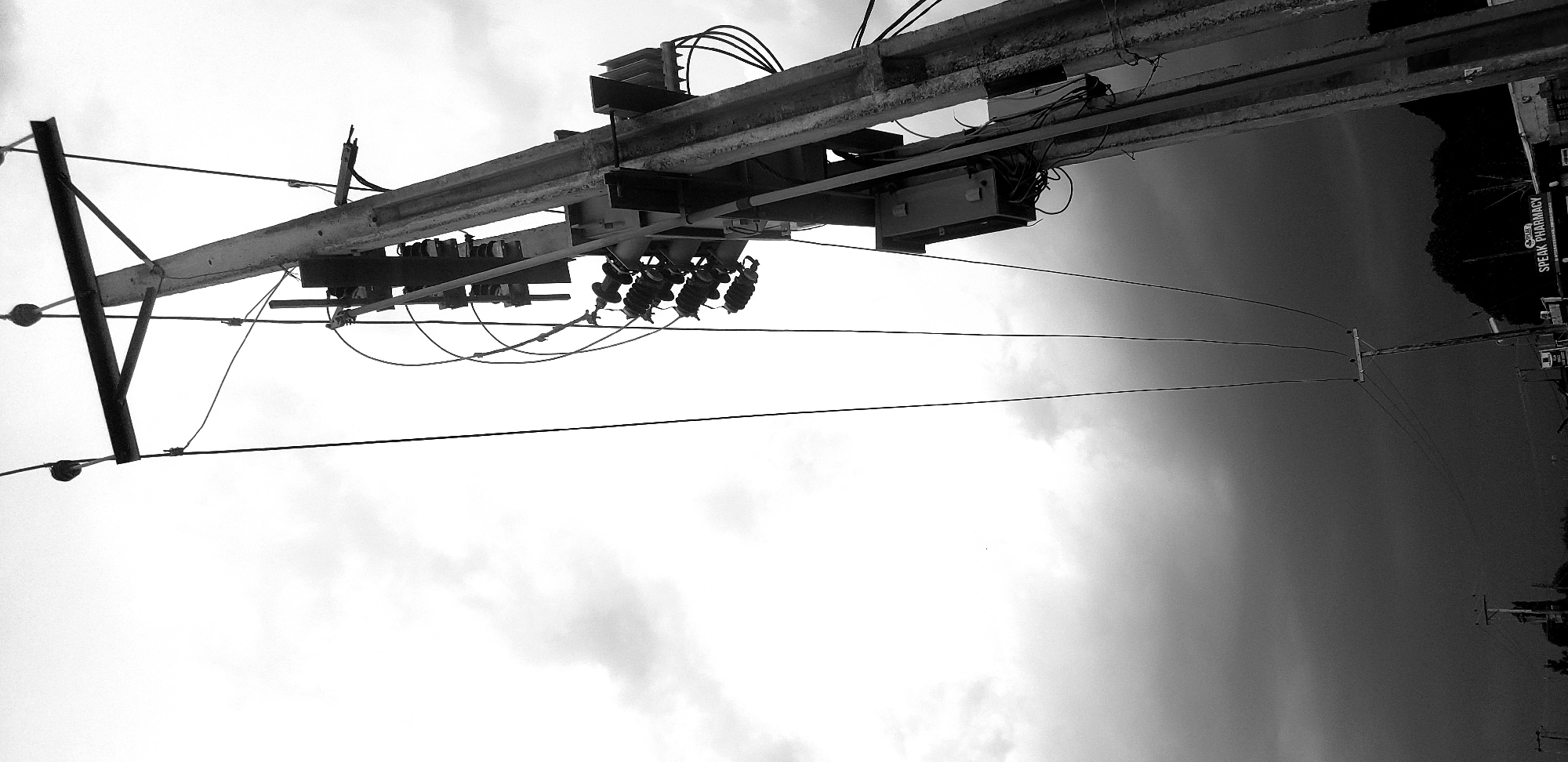 This black and white photograph, taken from a low angle, features a complex utility pole structure against a dramatic sky. A large, horizontal utility beam, equipped with various electrical components, boxes, and ceramic insulators, stretches across the upper part of the frame. Numerous wires and cables emanate from and connect to this structure, extending across the image. The monochrome palette emphasizes the textures of the metal and the intricate network of lines.
The sky dominates the background, displaying a stark contrast: bright, cloud-filled white and light grey on the left transition to a dark, heavily clouded or stormy grey and black on the right. This suggests significant cloud cover, possibly indicating an overcast day, or the approach of dawn or dusk. In the far distance, silhouetted against the dark sky on the right, appears to be a natural landscape, perhaps hills or dense foliage. Below this, a smaller, traditional utility pole is visible in the background, supporting additional lines.
No people are visible, and no specific event or activity is taking place; the scene captures a moment of static infrastructure and environment. A visible sign on the far right, against the dark background, clearly reads "SPEAK PHARMACY", with potentially a partial word like "CORES" above it, indicating a commercial establishment. The image, set in Narayi, Nigeria, focuses on the interplay of urban infrastructure and the natural elements of the sky and distant landscape.
