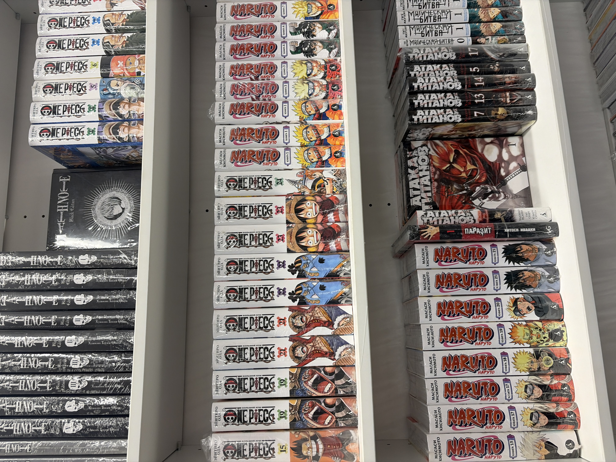 The image shows a close-up of three shelves, likely in a retail store, stocked with manga. From left to right, the first shelf contains multiple copies of "Death Note Black Edition," the second shelf contains copies of "One Piece" and "Naruto", and the third shelf contains copies of "Jujutsu Kaisen", "Attack on Titan", "Parasyte", and "Naruto". All manga titles appear to be translated into Russian. The spines of the books are visible, with their titles clearly displayed. The books are packaged and appear to be new. The setting is unspecified.