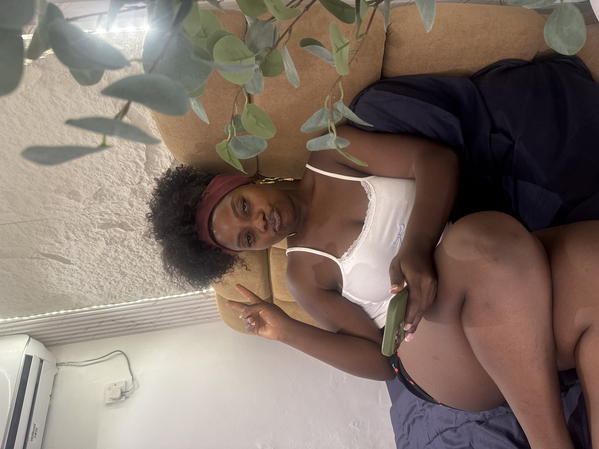 A woman with dark skin and curly black hair, wearing a maroon headband, a white tank top, and black shorts with a cherry pattern, is sitting on a brown sofa. She is holding a green smartphone in her right hand and has her left hand raised in a peace sign. She is wearing gold dangle earrings. The background consists of a textured white wall with vertical white strips and an air conditioning unit mounted high on the wall. A potted plant with green leaves is to the right of the woman. The scene appears to be indoors, possibly in a bedroom or living room, and could be located in Abuja, Nigeria, given the context of the question.