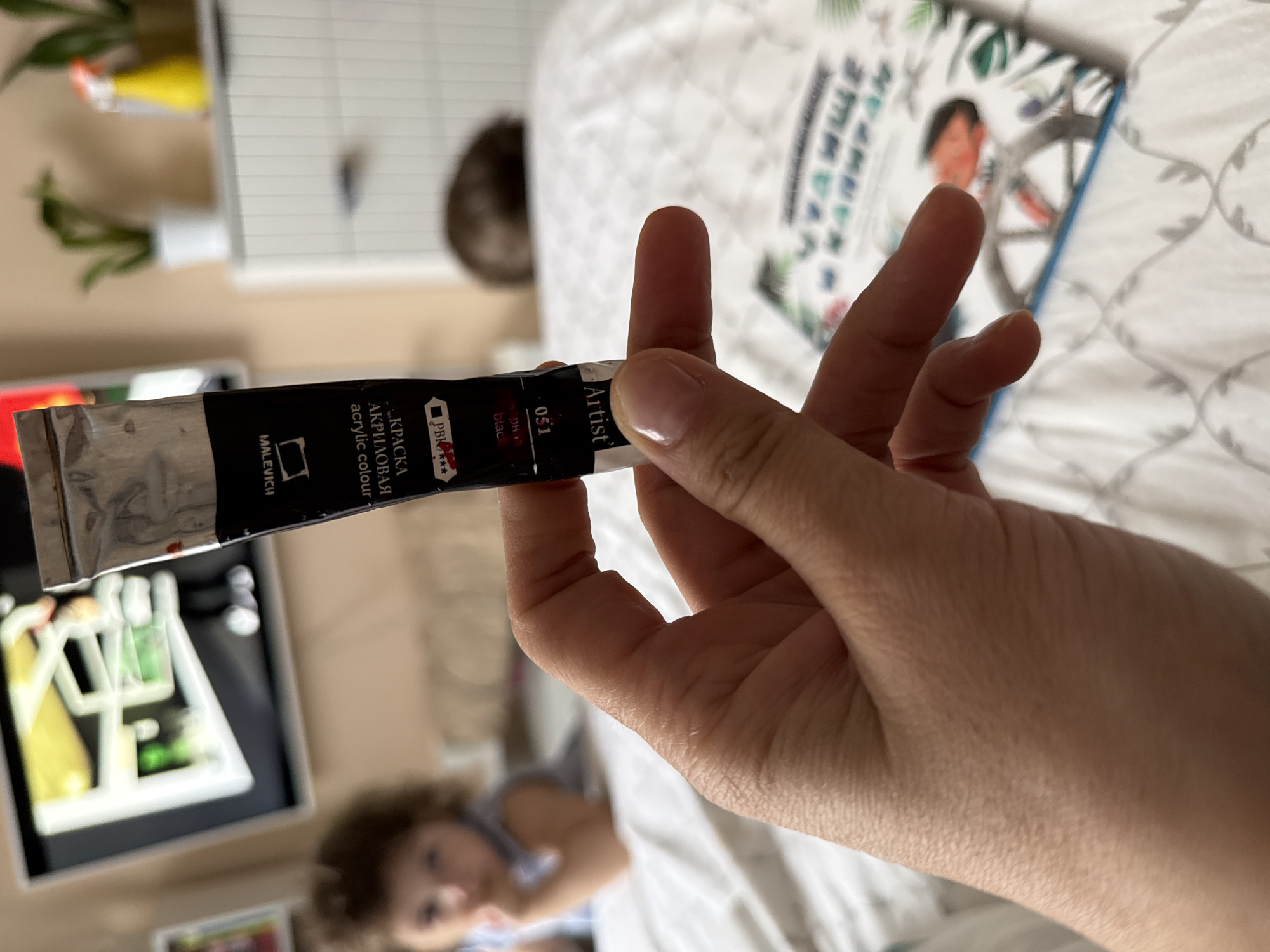 A person's hand holds a black tube of "Malevich" artist acrylic paint in front of a patterned white bedspread. The paint tube label includes the words "acrylic colour" and "КРАСКА АКРИЛОВАЯ". The label also indicates "051" and "PBr ***". In the background, a blurred image of a child is visible, as well as a book with an illustration. In the far background, a shelf with plants and a painting are visible. Tobolsk, Russia is not mentioned in the image.