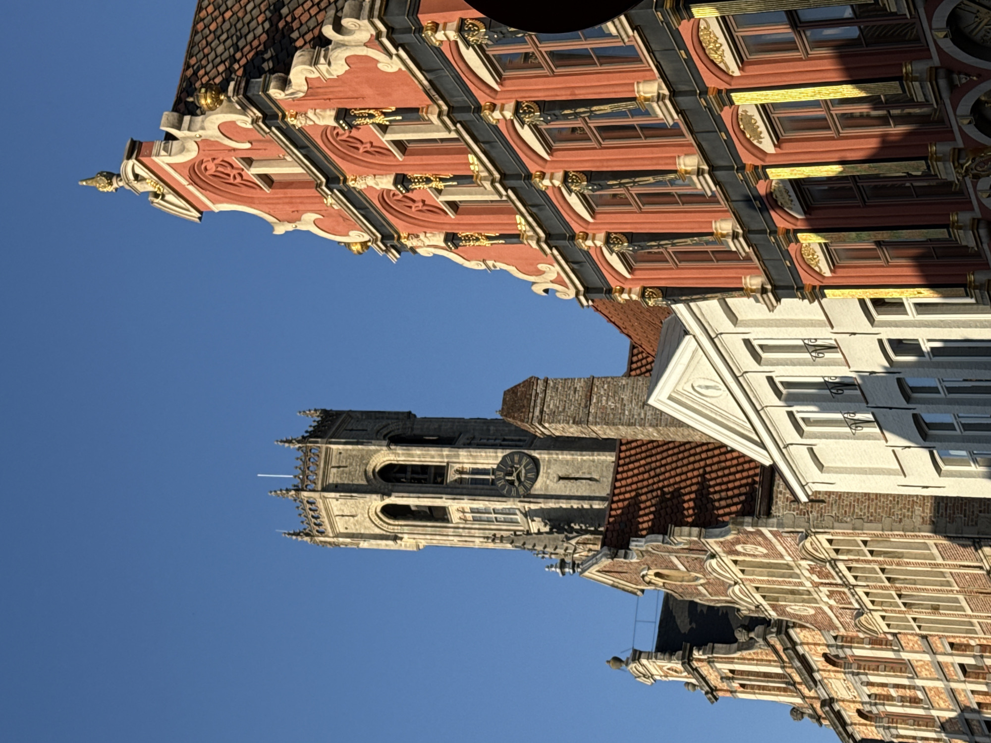 The image depicts a street scene in Bruges, Belgium, with a clear blue sky as the backdrop. In the foreground, several historic buildings with distinct architectural styles are visible. To the right, a predominantly red building with ornate gold detailing and numerous windows stands out. Its facade features a stepped gable with decorative elements and a tiled roof. Adjacent to it, a white building with a simple, clean facade and windows is situated. Further to the left, a brick building with a pitched tiled roof and multiple windows is shown. Dominating the background is the Belfry of Bruges, a medieval bell tower constructed of grey stone. The tower has a clock face and arched windows, topped with a decorative spire and battlements. The lighting suggests it is either early morning or late afternoon, casting shadows across the facades of the buildings. No people are visible in the image.