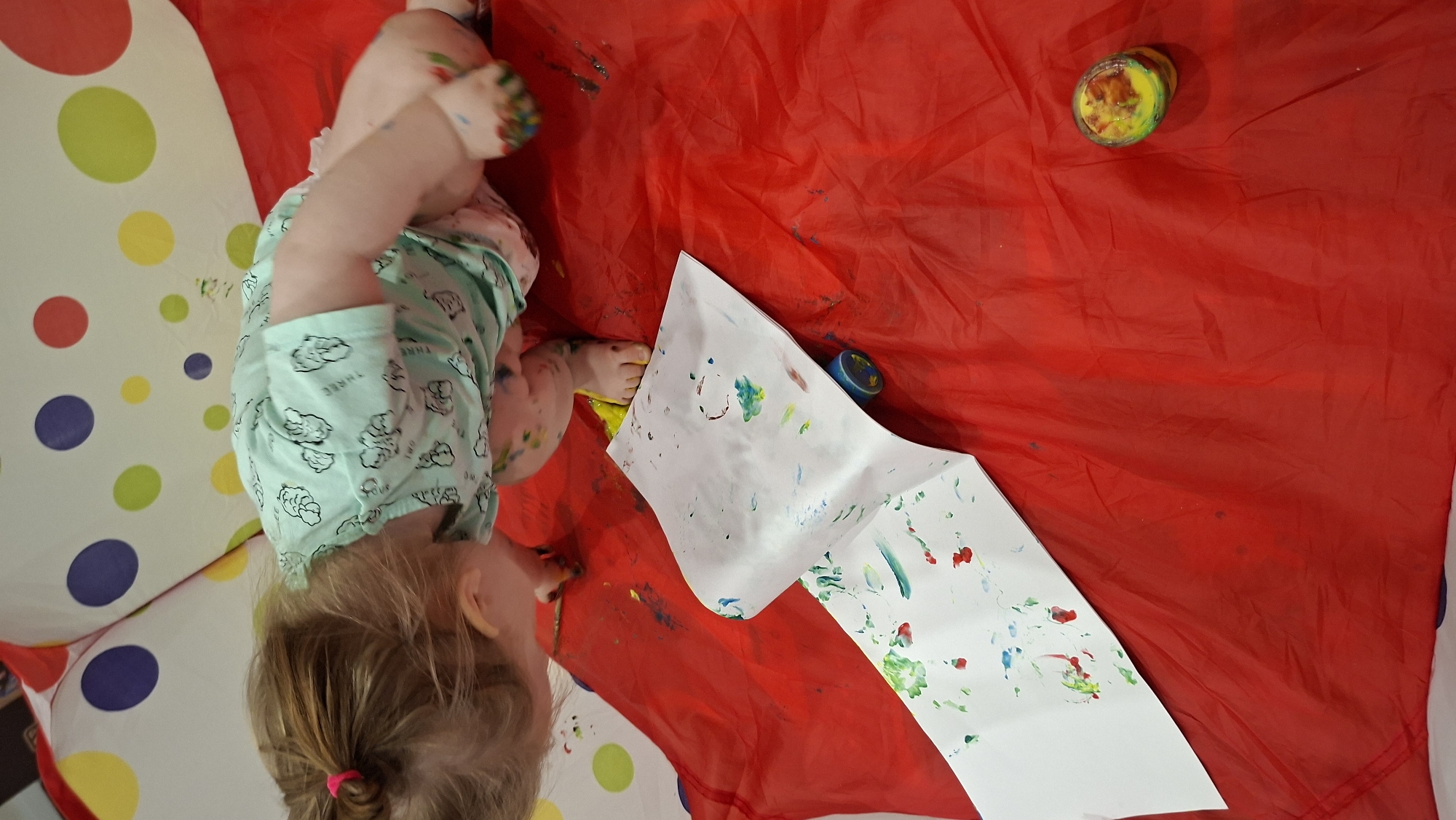 The image shows a child lying on a red surface with a large sheet of white paper. The child is wearing a light green shirt and has blonde hair with a pink hair tie. The child's hands and feet are covered in paint. There are two small containers of paint nearby. The background includes a surface with large polka dots. Based on the prompt, this activity is taking place in Chaikovsky, Russia.
