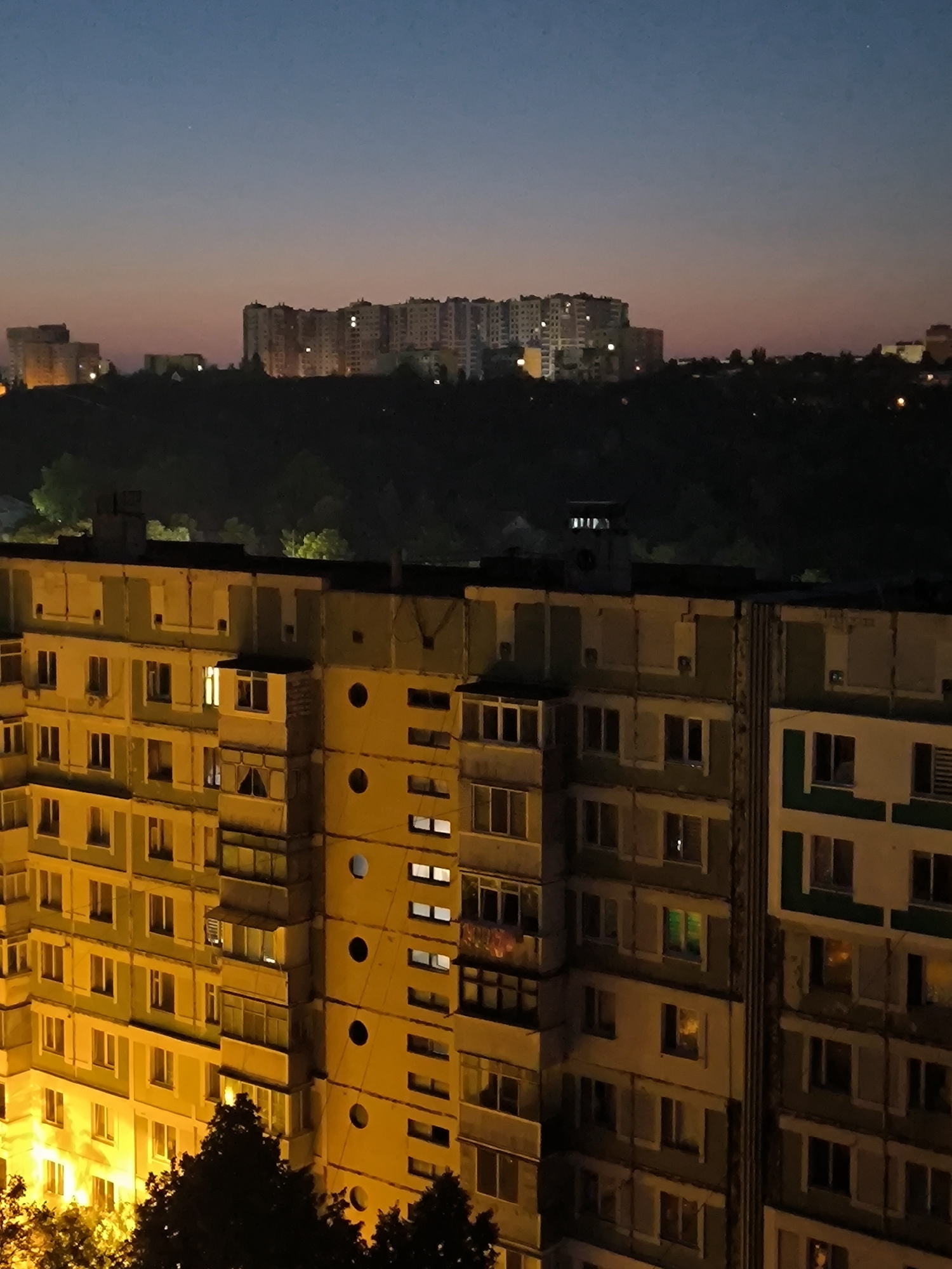 The image captures a twilight view of apartment buildings in Chișinău, Moldova. The sky transitions from a deep blue at the top to hues of pink and orange near the horizon, indicating the end of the day. In the distance, silhouetted against the fading light, are rows of multi-story apartment blocks, some with illuminated windows.
In the foreground, two prominent apartment buildings dominate the scene. The building on the left is a tall, light yellow structure with numerous windows, many of which are lit, casting a warm glow. The building on the right appears to be a similar height, with sections of light yellow and light green facades. This building also features many windows, with some illuminated by interior lights, displaying a variety of colors, including green and orange.
The architecture of the buildings suggests a Soviet-era design, characterized by repetitive window patterns and utilitarian construction. Trees and foliage are visible in the lower parts of the image, some illuminated by the light from the buildings, adding a touch of nature to the urban landscape. The overall atmosphere is quiet and still, characteristic of the transition from day to night in a residential area. The image is a snapshot of urban life at dusk, showcasing the interplay of natural light and artificial illumination on the city's residential structures.
