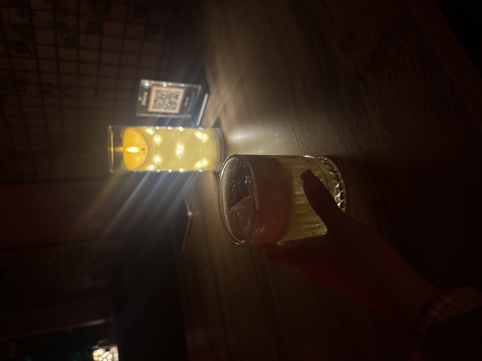 A slightly low-angle shot of a hand holding a glass of a light-colored drink with ice, sitting on a dark wooden surface. Next to the glass, there's a glass container filled with a yellow liquid and decorated with small lights to resemble a candle, and a menu stand with a QR code. The environment appears to be a dimly lit bar or restaurant, likely in Batumi, Georgia. The overall lighting suggests a warm, cozy atmosphere. The hand is holding the drink as if about to take a sip or show it off.