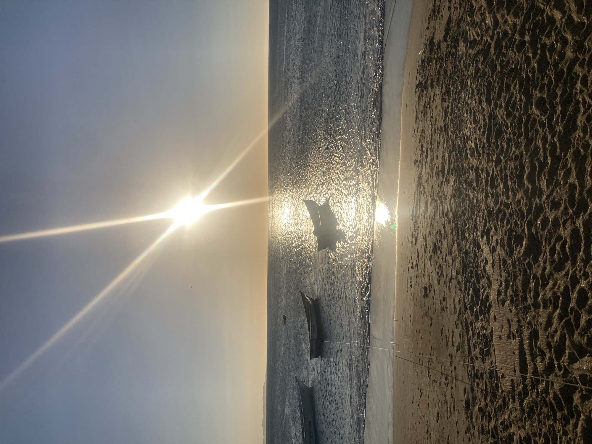 This photograph shows the beach at Juan Griego, Venezuela, during sunset. The sun is positioned in the upper left of the frame, producing intense glare and rays across the scene. The ocean occupies the central part of the image, its surface reflecting the sunlight. Several fishing boats are in the water, some appearing to be tied to the shore with ropes. The beach takes up the right side of the image, its sand dark and textured. There are no people visible.