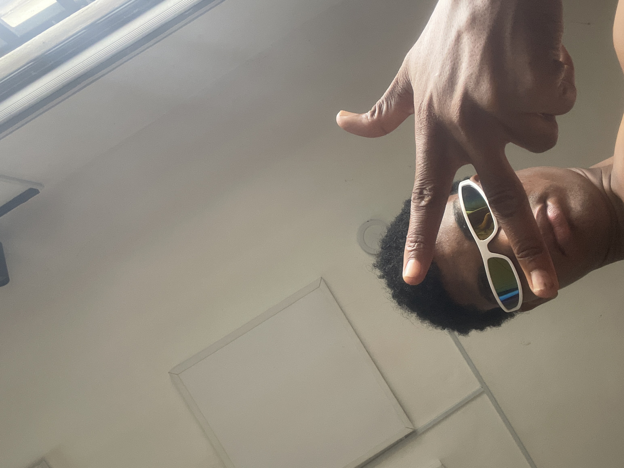 The image shows a close-up, low-angle shot of a young Black person wearing white-framed sunglasses with mirrored lenses. Their right hand is positioned in front of their face, with fingers extended, obscuring part of their face and the sunglasses. The background is a plain, light-colored wall with what appears to be a ceiling light fixture and possibly a window or light source to the right. The context of "Ado, Nigeria" is not visually ascertainable from the image itself.