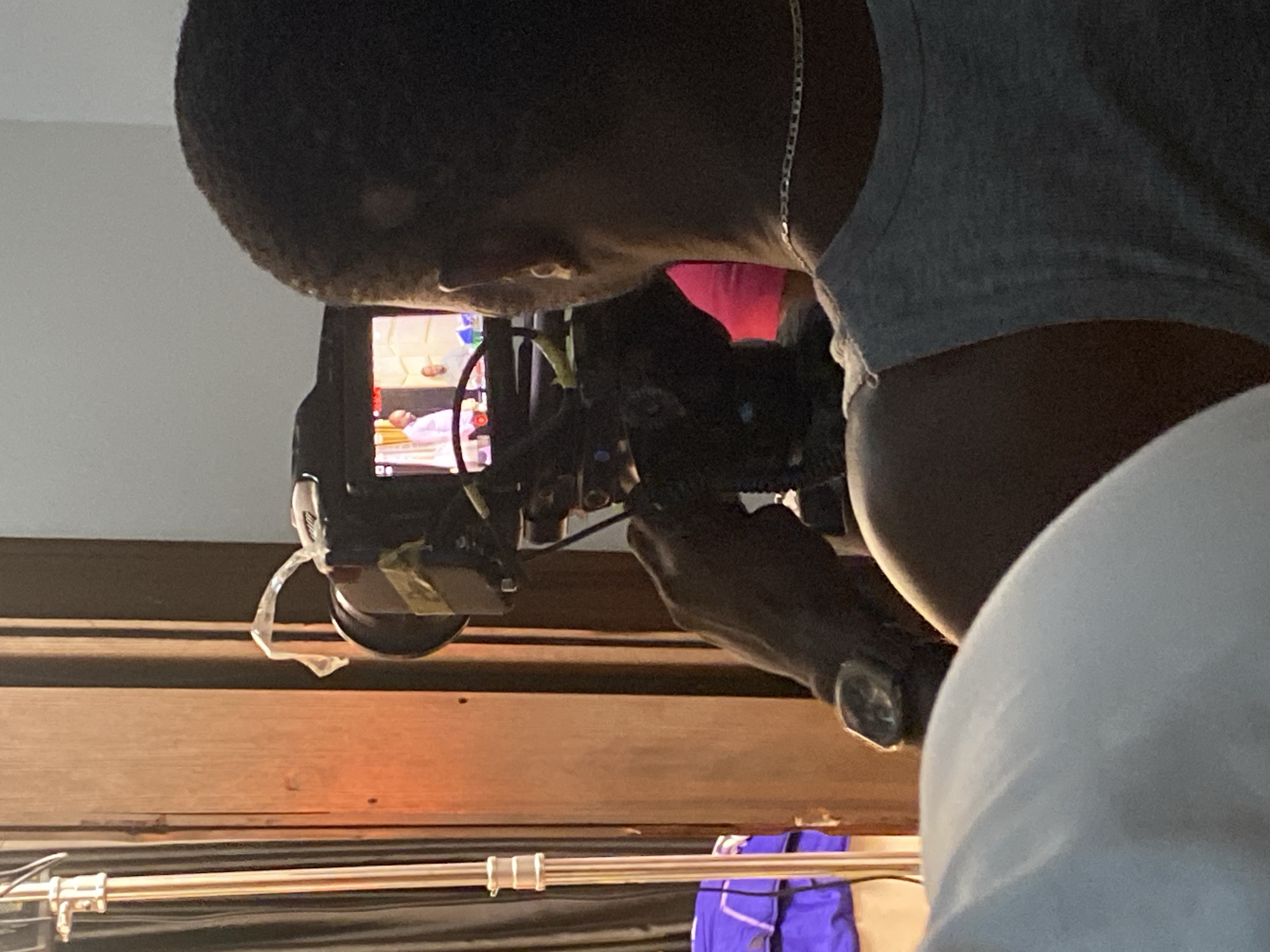 A dark-skinned individual, seen from the upper back and right side, is actively operating a black digital camera. The person has short, dark hair and is wearing a grey t-shirt and a silver chain necklace. Their right hand is visible holding the camera.
The camera's articulating screen displays an image of an indoor scene, where a person appears to be lying down, possibly on a bed or table, with what looks like medical or patient care equipment nearby, including a white sheet/gown and a blue/white striped object that could be part of an IV stand or monitor. Another person's head is partially visible in the upper right of the screen's frame. A small, unidentifiable white logo or text is on the camera body above the screen. A piece of clear tape or plastic hangs from the camera's lens or body.
The setting appears to be indoors, potentially a film set or a professional shooting location. A prominent dark wooden beam or panel runs horizontally in the mid-ground. Above this, the background is a plain, light-colored wall. Below the wooden structure, a silver bar, likely part of a light stand or equipment rig, spans horizontally across the foreground. Behind this bar and below the wood, a vibrant blue fabric object, possibly a bag or cloth, is visible. There is a distinct red or pink light glowing in the shadowy area behind the operator's neck. The lighting suggests an artificially lit environment. The person's posture indicates focused engagement with the camera. No discernible text is clearly readable in the main image.