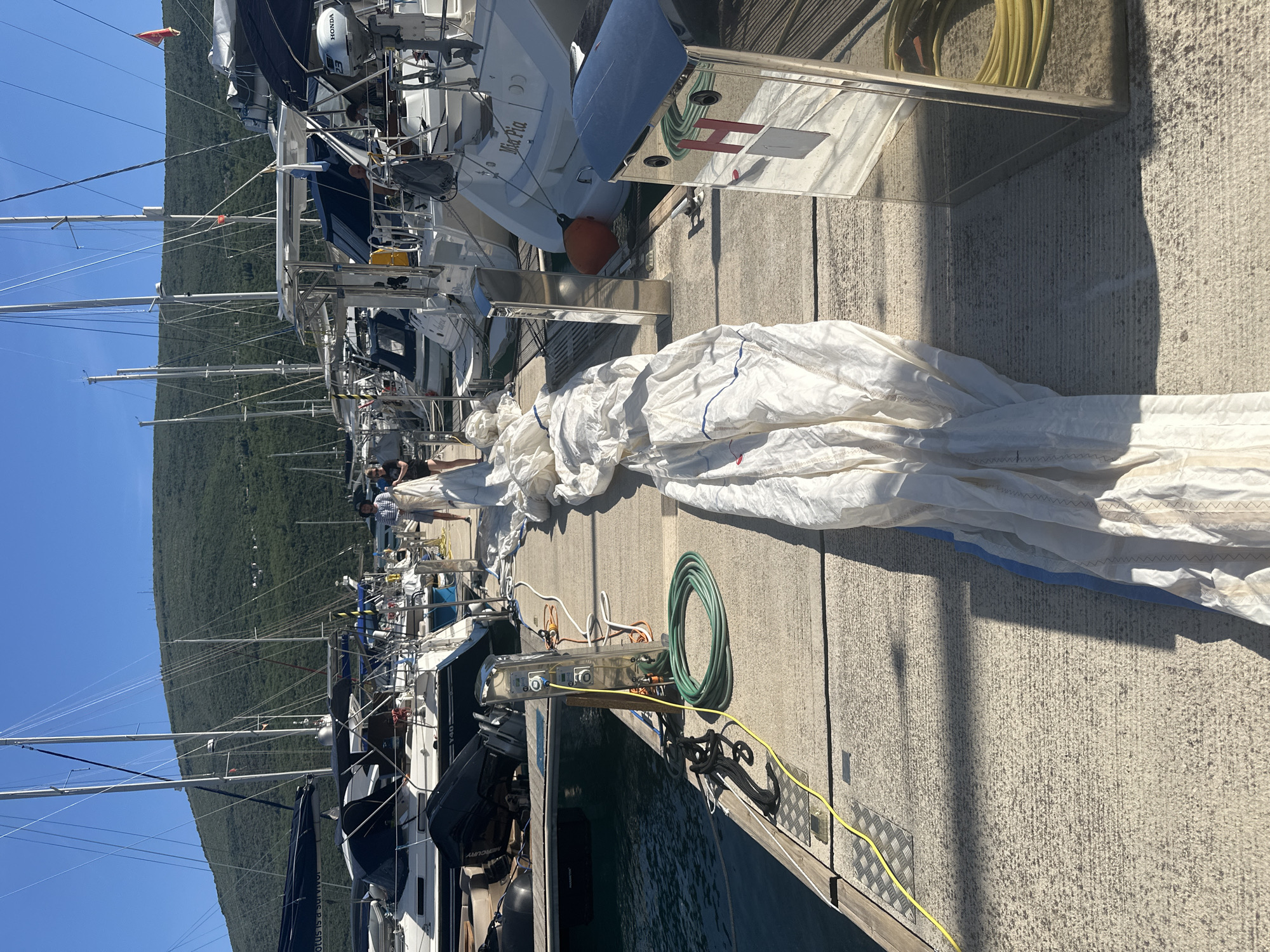 The image captures a marina scene in Herceg Novi, Montenegro, under clear blue skies and bright sunlight. A concrete dock, textured with a ribbed surface and featuring sections of metal diamond plate, extends into the foreground and middle ground. Along the right side of the dock, a large white sail, possibly a genoa or mainsail, is laid out, partially folded and extending longitudinally. Two individuals are positioned further down the dock, near the sail. One person, wearing a dark top and light shorts, stands on the left side of the dock near the boats. Another individual, dressed in a striped shirt and dark shorts, appears to be actively handling the sail. A third person's lower legs are visible on the far right, also near the sail.
Multiple boats are moored alongside the dock. Predominantly sailing yachts with visible masts and rigging line the berths. One white sailing yacht in the middle ground has "Mia Pia" inscribed on its stern. Another boat on the left side features "Y40" and "Lotus is a survivor" text. Outboard engines, including brands like Mercury and Honda, are mounted on some vessels. Various nautical equipment, such as black coiled mooring lines, a coiled green garden hose, and a yellow electrical power cable, are visible on the dock. Chrome-finished utility pedestals, equipped with electrical outlets and water spigots, are spaced along the dock; one prominent pedestal displays a red "H" symbol. In the background, a large, lush green mountain range dominates the horizon, with scattered small white structures visible on its slopes, all set against a clear, sunny sky.