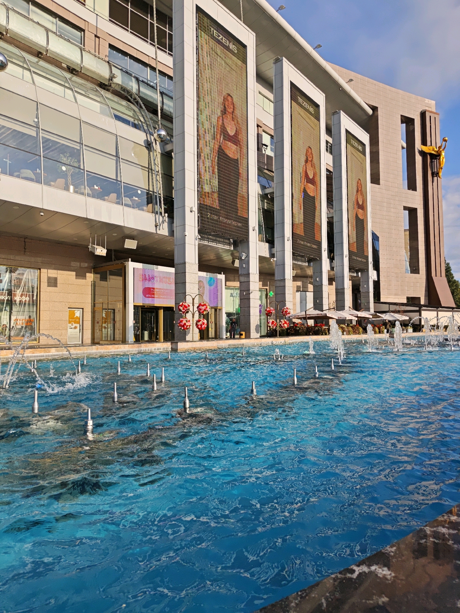 The image depicts a vibrant urban scene in Moscow, Russia, likely at the Tezenis store located in the shopping mall "European" (Evropeyskiy). In the foreground, a large, blue fountain with multiple jets of water creates a dynamic display. The water is bright and appears well-maintained.
Behind the fountain, the facade of a modern building is visible, characterized by a combination of glass and beige stone. Large, vertical digital billboards are prominently displayed on the building's exterior. These billboards feature advertisements for Tezenis, a clothing brand, with images of models wearing athletic apparel. The building's architecture includes large glass windows, suggesting shops or restaurants on the lower levels, and balconies or upper floors with seating areas visible through the glass.
Several umbrellas and outdoor seating are set up in front of a cafe or restaurant area. The scene is bathed in natural light, indicating it is daytime. A golden statue of an angel or winged figure is mounted on the side of the building on the right, adding an artistic element to the architecture. The sky is visible in the background, with some clouds, suggesting clear weather. There are no people clearly visible in the immediate foreground, though a few figures may be present in the distance, near the entrance of the building and in the outdoor seating area. The overall impression is one of a lively, contemporary urban environment.