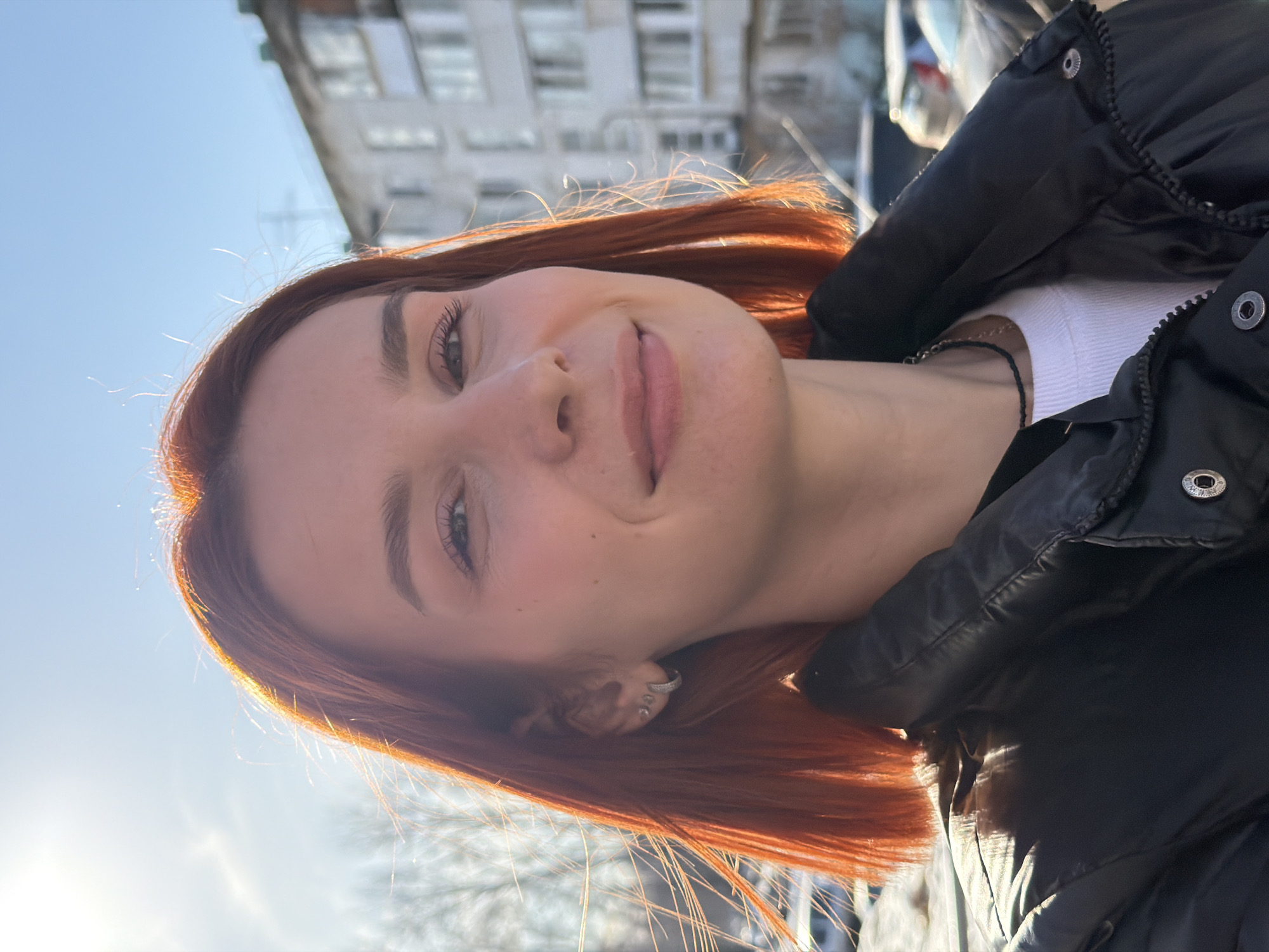 A young adult female with short, straight, reddish-orange hair is captured in a close-up, chest-up shot. She faces the camera directly with a slight, closed-mouth smile. Her visible left ear is adorned with a small, stone-studded hoop earring and at least two small stud earrings. She wears a black puffer jacket over a white ribbed top, accessorized with a black choker necklace. The scene is outdoors under a clear blue sky with bright sunlight from behind and to the left of the subject, creating backlighting on her hair and a lens flare in the upper left quadrant. In the background on the right, a multi-story residential building with numerous windows is visible. To the left, bare tree branches are discernible, and indistinct vehicles are partially visible further in the background. The depicted location is an urban outdoor setting within the city of Chaikovsky, Russia.