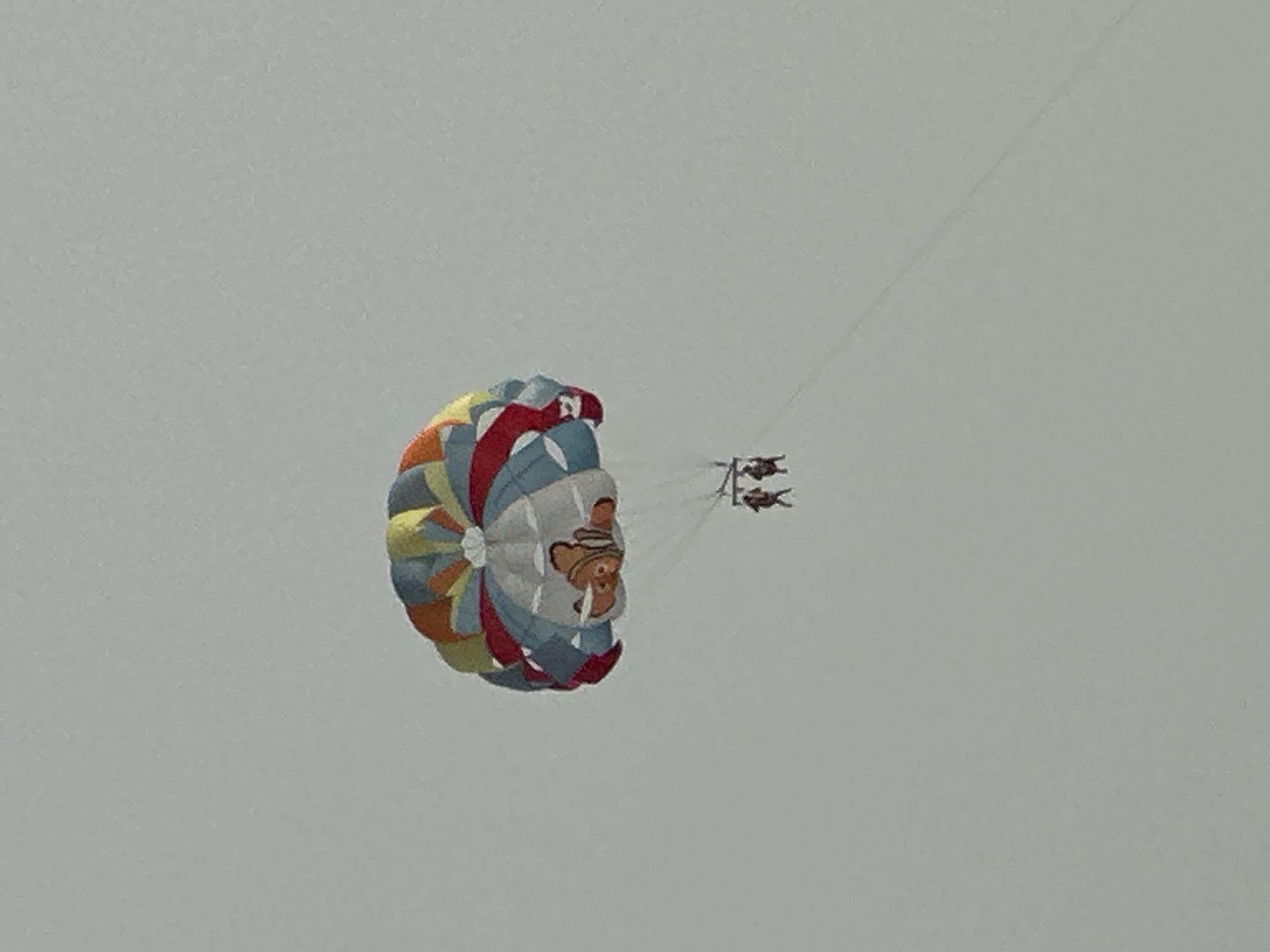 In Rafailovići, Montenegro, two individuals are parasailing under a colorful parachute displaying a character resembling Nemo from Finding Nemo. The parachute is predominantly white with sections of blue, red, yellow, and orange. The two individuals are suspended below the parachute, appearing to be attached by harnesses and ropes. The background consists of a light gray sky, indicating an overcast or hazy day. A thin rope or tow line extends diagonally from the parachute down towards the lower right of the frame, suggesting the activity is being conducted by a boat or other towing mechanism out of frame. The overall scene captures a recreational activity taking place in an outdoor, airborne environment.
