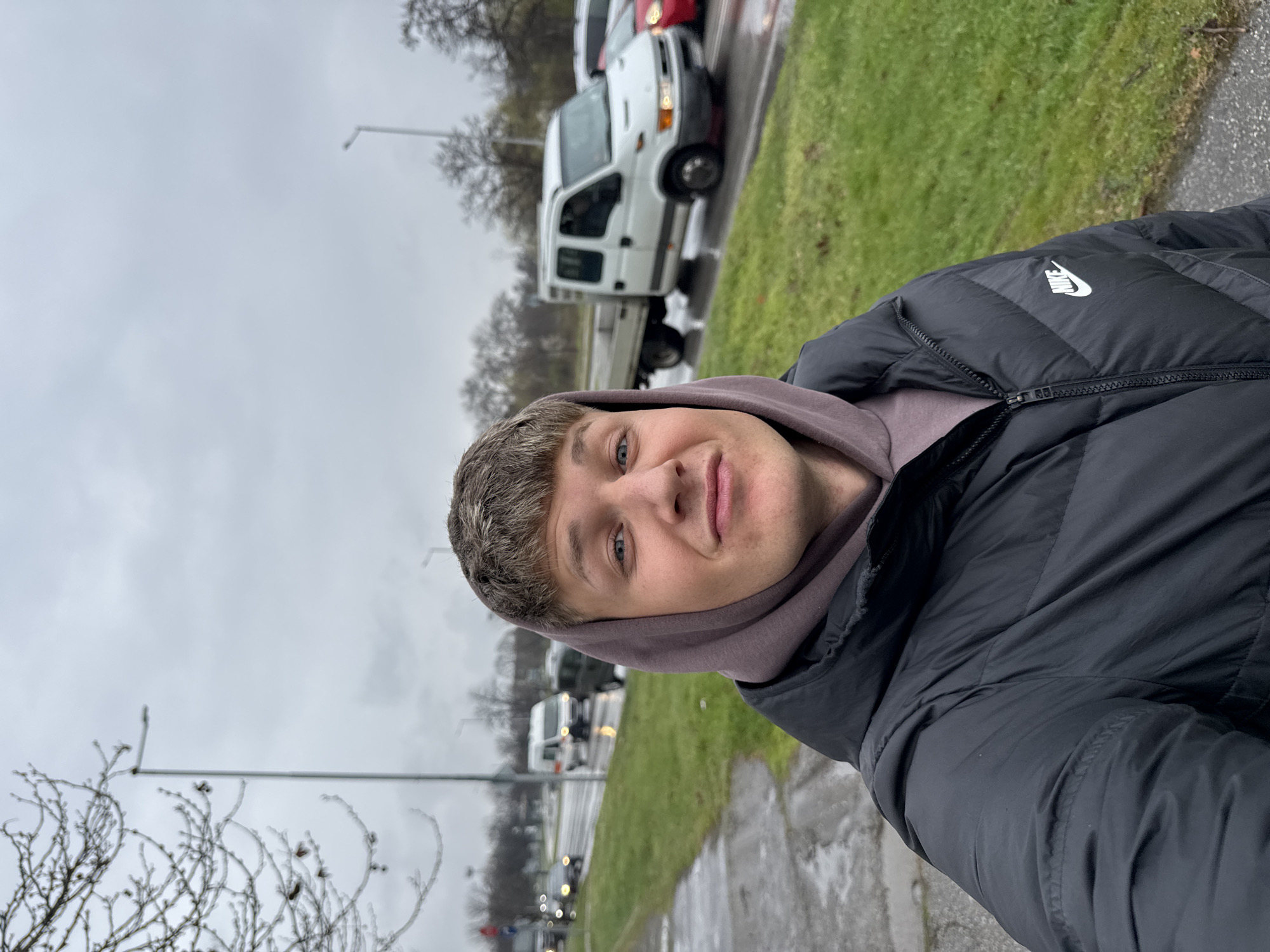A man in a black Nike puffer jacket and light purple hoodie is taking a selfie. He has short brown hair and blue eyes. The background shows a cloudy day with a street, green grass, bare trees, street lights, and cars, including a white truck. The location appears to be Plovdiv, Bulgaria.