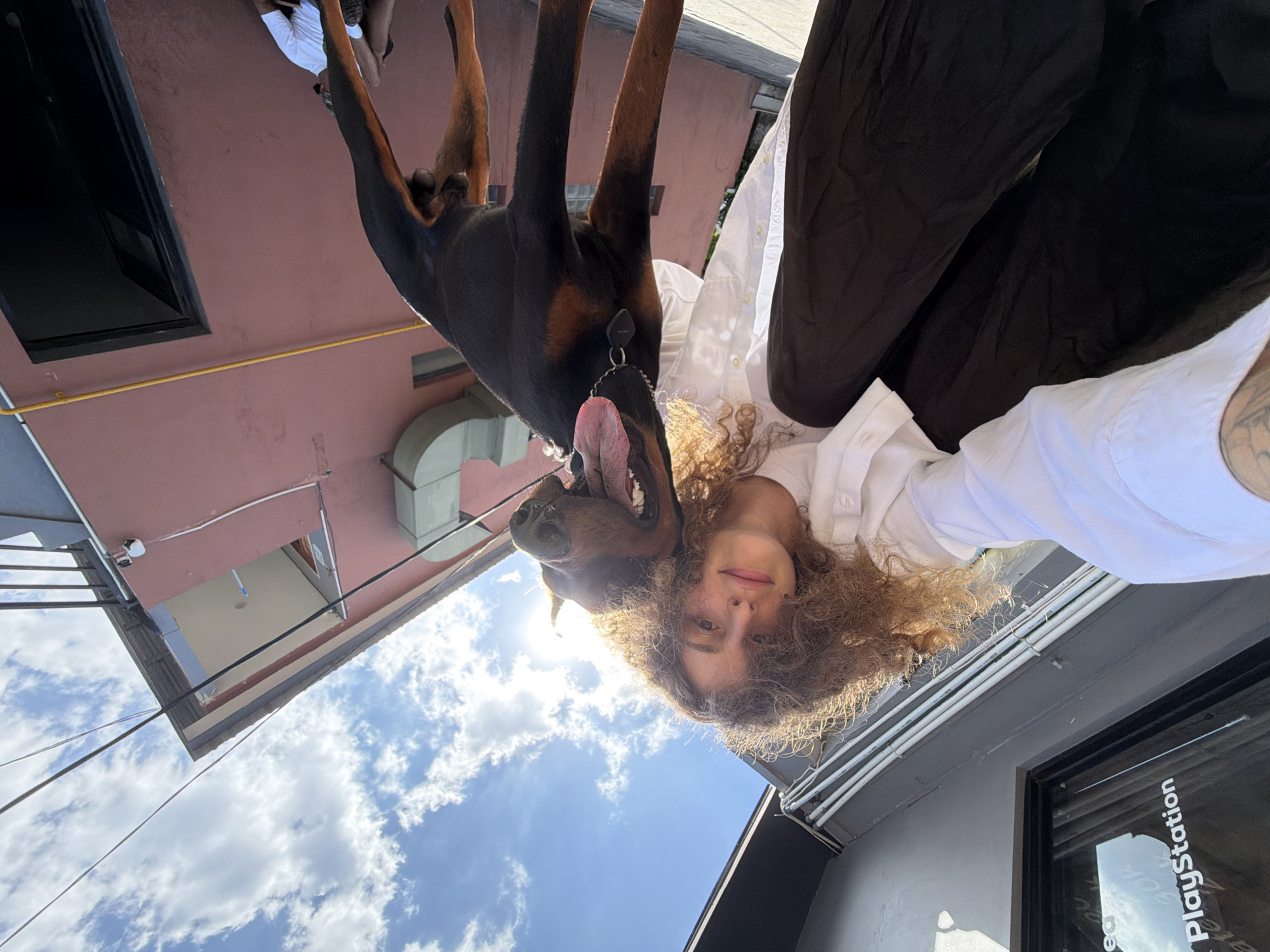 A low-angle, wide-shot photograph captures a person with curly brown hair and a Doberman pinscher in front of a salmon-colored building. The person, wearing a white button-up shirt and dark pants, is squatting and holding a phone to take a selfie. The Doberman, facing the camera with its tongue out, is positioned in front of and slightly to the right of the person.
The building's exterior features a dark-framed window on the left, reflecting the sky and clouds, with "PlayStation" text visible. The building's facade has visible piping and a large "C" shaped structure on the upper left. A dark doorway is visible on the lower right. In the lower right background, a person in a white shirt and dark shorts is seated. The scene is outdoors under a bright, partly cloudy sky, with power lines crossing the frame. The overall lighting suggests daytime. This scene is located in North Kuta, Indonesia.