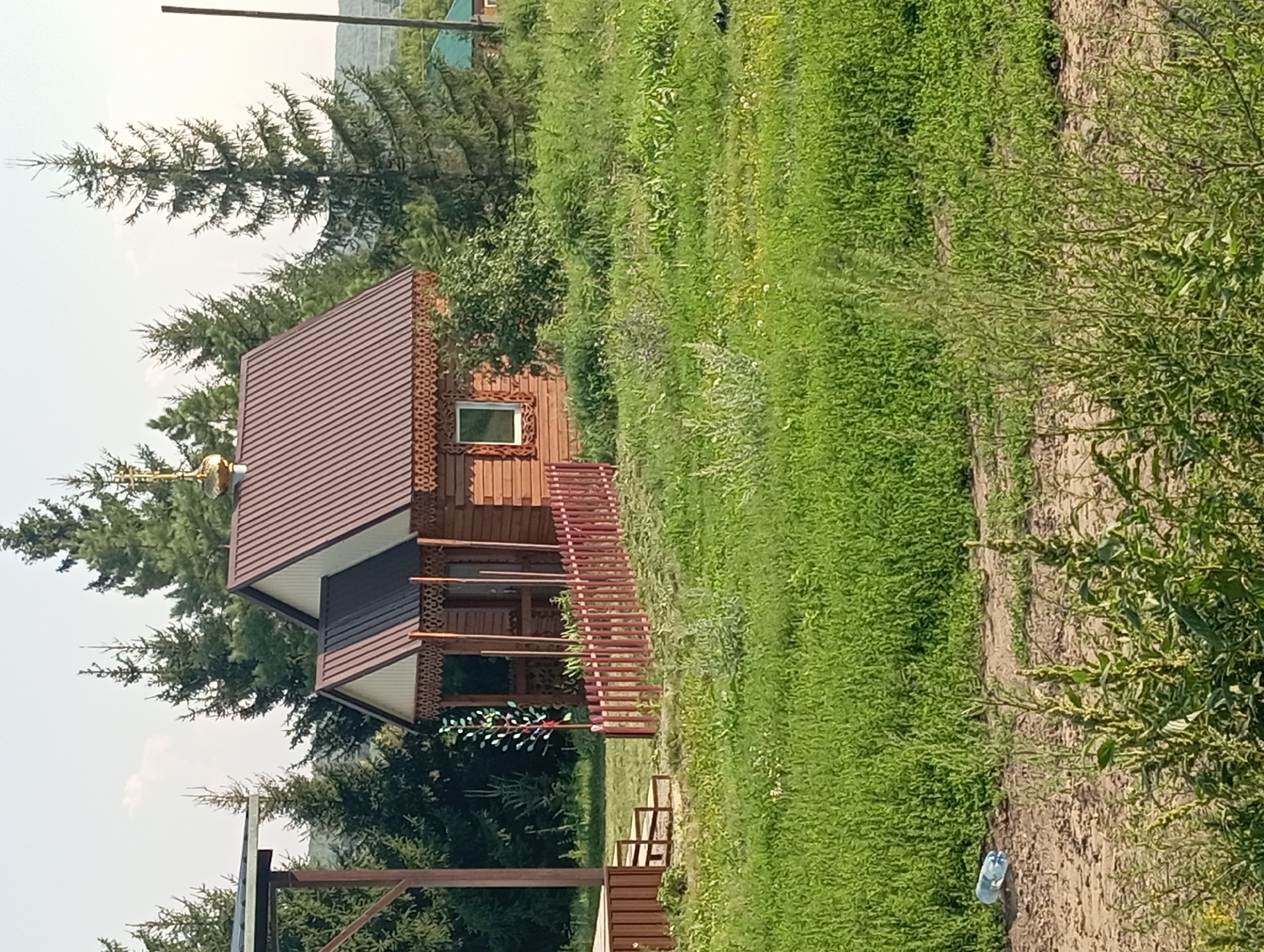 The image depicts a small wooden building with a brown metal roof, possibly a chapel or a miniature house, situated in a rural setting. The building features a decorative wooden trim under the eaves and a single window. A small red wooden fence encloses a portion of the property in front of the building, and a metal sculpture resembling a tree with colorful leaves stands near the fence. The surrounding environment is characterized by lush green grass and several tall evergreen trees. In the foreground, a dirt path is visible with a discarded plastic water bottle. In the background, distant hills and other structures with green roofs can be seen. The scene appears to be in Новокопылово, Russia, given the context provided. There are no people present in the image.