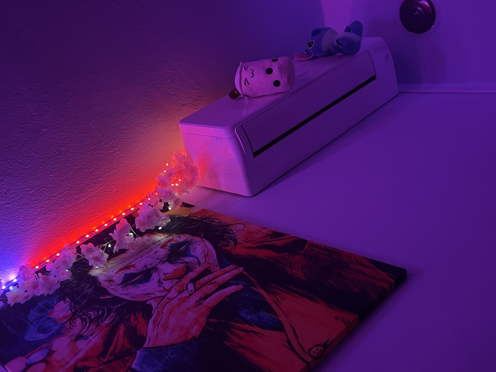 This image captures an interior scene within a room located in Debelt, Bulgaria, illuminated primarily by a deep purple ambient light with additional red and blue accents. A large canvas art piece depicting a stylized rendition of the Joker character from the film *Joker* dominates the left side, showing the character with a hand partially obscuring his face. Above this artwork and extending across the top right wall is a strip of red LED lights, intertwined with a decorative garland of artificial white or light pink cherry blossom-like flowers. Mounted high on the right wall is a white ductless mini-split air conditioning unit. Positioned on the top surface of the air conditioning unit are two plush toys: a light pink, smiling bubble tea cup plush with black boba dots, and partially visible below it, a blue plush figure of the character Stitch. The walls exhibit a textured finish. In the lower right corner, a dark, circular fixture is affixed to the wall.