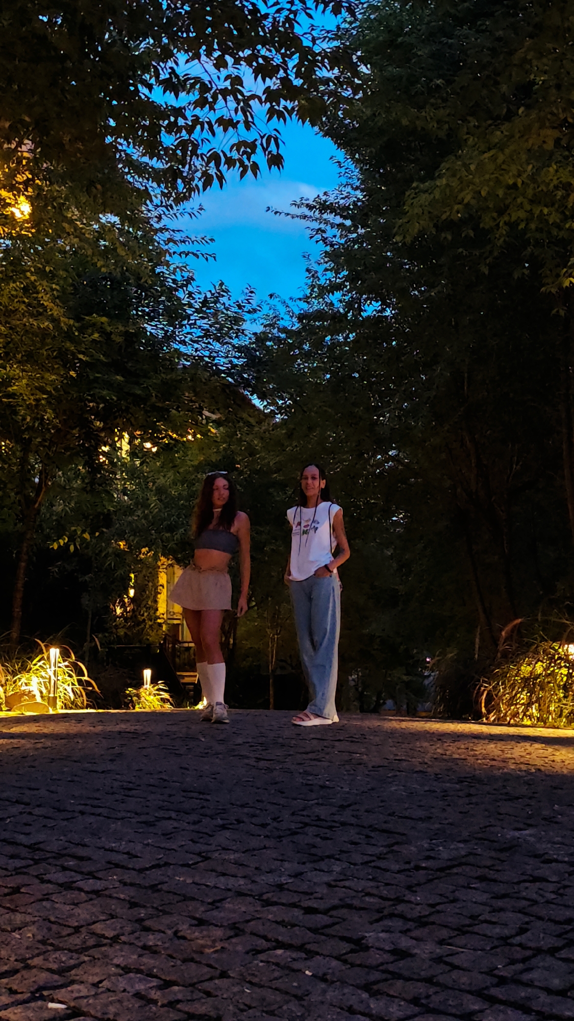 The image features two young women standing on a cobblestone path in an outdoor setting at dusk or early night. Lush green trees and foliage form a natural archway overhead, with a deep blue sky visible through the canopy. Warm light from ground-level lamps illuminates the surrounding plants and portions of the path.
The woman on the left wears a gray strapless crop top, a light brown mini skirt, white knee-high socks, and light-colored sneakers. Her long, dark hair flows over her shoulders, and she has sunglasses perched on her head. She stands with a relaxed posture.
The woman on the right is dressed in a white t-shirt featuring a colorful graphic design with some indistinct text, paired with light blue wide-leg jeans and light-colored slip-on shoes. Her dark hair is pulled back, and she also has glasses on her head. Her hands are in her pockets, and she displays a calm expression.
Both women are looking towards the camera, appearing to pose casually for a photograph. The scene conveys a tranquil atmosphere, characteristic of an evening in a park or garden. The overall setting, combined with the artificial lighting, suggests a pleasant, well-maintained outdoor space in Púng Luông, Vietnam.