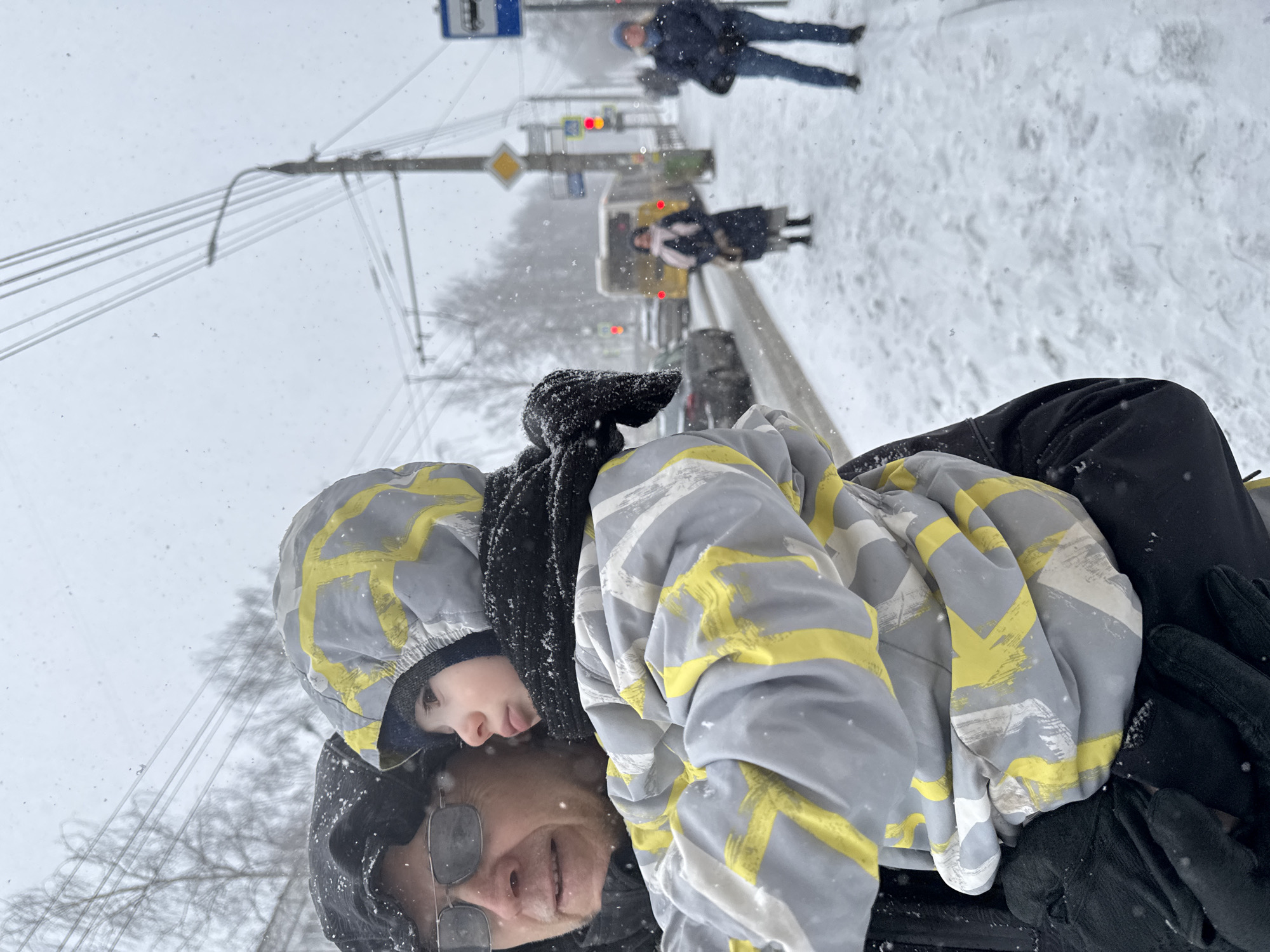 In the city of Izhevsk, Russia, an older individual wearing glasses and a black hooded jacket is holding a child bundled in a grey and yellow jacket with a black scarf. They are standing on a snow-covered street, with other pedestrians visible in the background. Streetlights, a bus stop sign, a crosswalk sign, and a traffic light are present, indicating an urban environment. The traffic light appears to be showing red. Snowflakes are falling.