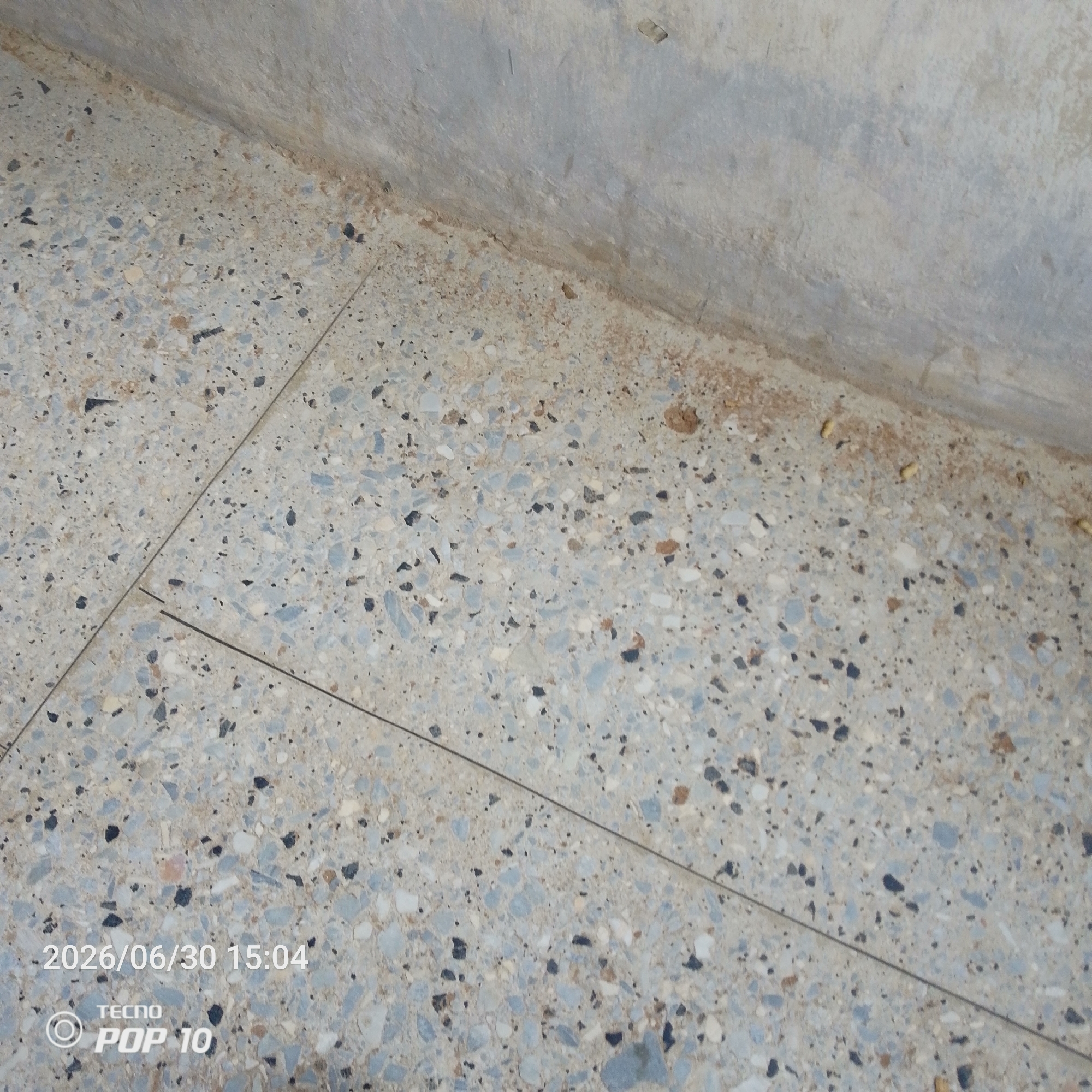 The image shows a close-up view of a terrazzo floor meeting a concrete wall. The floor is composed of small chips of various colors, including gray, white, blue, and black, embedded in a lighter gray matrix. Two faint lines, likely grout lines or cracks, run diagonally across the floor. The wall is textured and appears to be made of concrete. Along the base of the wall, there is a strip of dirt and debris.
In the lower left corner of the image, text is visible. It reads "2026/06/30 15:04" followed by "TECNO POP 10". This indicates the date and time the photo was taken, as well as the brand and model of the phone used.
The scene appears to be indoors or in a covered outdoor area, given the presence of the wall and the nature of the flooring. The lighting suggests it is daytime, likely in the afternoon based on the timestamp. There are no people or animals visible in the image, and the overall mood is neutral and observational, focusing on the texture and details of the floor and wall. The location context of Jalingo, Nigeria, does not offer specific visual cues within this image itself.