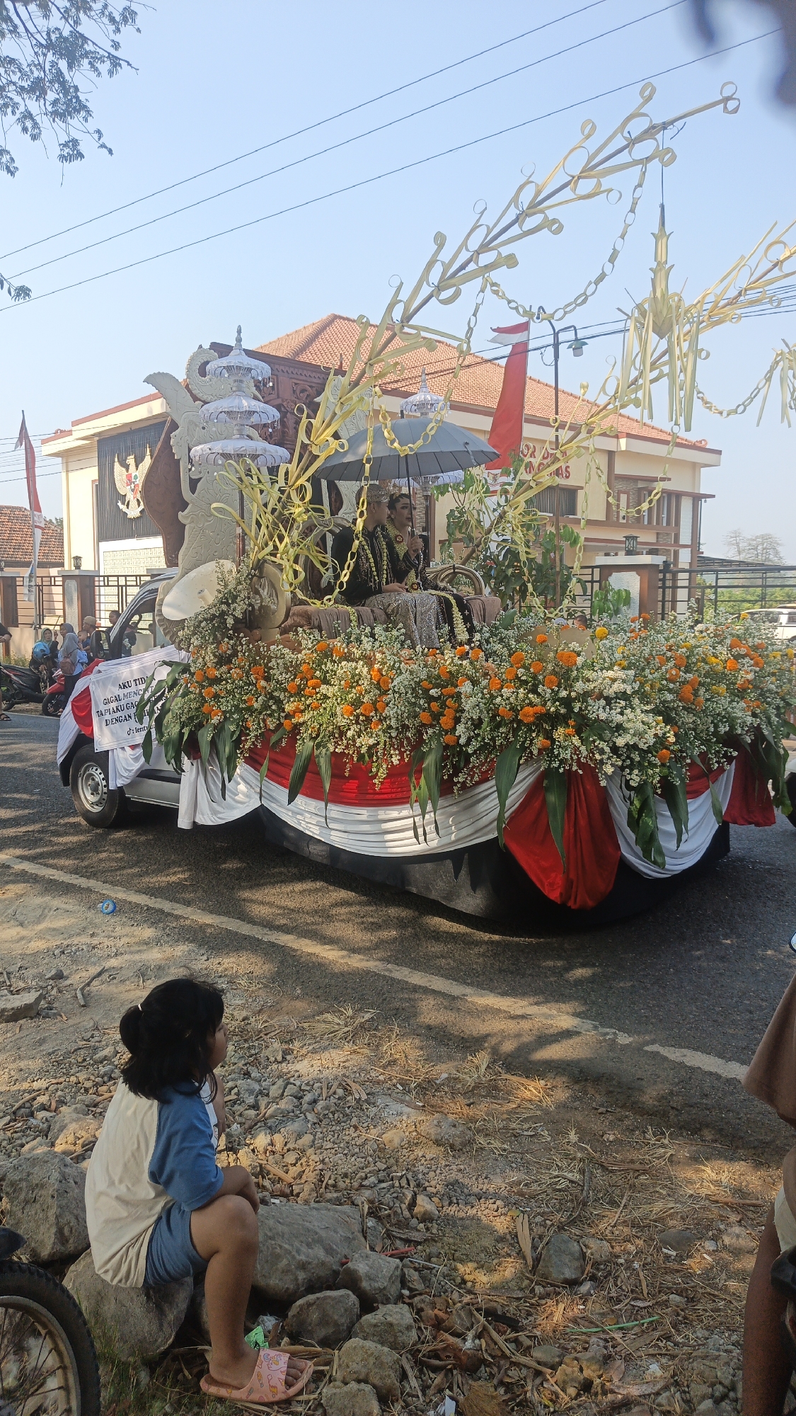 In the city of Kandangmas, Indonesia, a parade float is captured during daytime. The float, adorned with an abundance of orange and white flowers, is mounted on a small vehicle. Two individuals, dressed in traditional attire and wearing crowns, are seated on the float, holding an umbrella. The float also features decorative elements made of palm leaves and chain-like structures. In the foreground, a young girl with her hair tied back sits on rocks, facing away from the camera. The background reveals a building with a tiled roof, a national flag, and a clear sky. The street is paved, with some areas appearing unkempt and containing debris. Several other individuals and parked motorcycles are visible in the background.