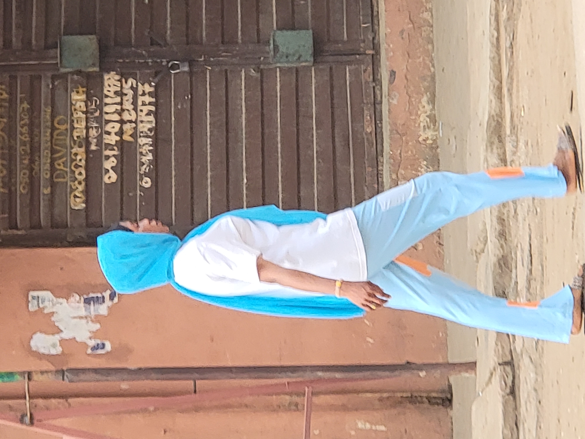 A person in a light blue hooded tracksuit and white t-shirt stands outside a building in what is likely the city of Maiburiji, Nigeria. The person is wearing brown shoes. The building has a brown corrugated metal door or gate with green metal squares at the top. Writing is visible on the metal door in yellow paint. The ground is a mixture of brown earth and pavement. Patches of peeled paint are visible on the adjacent walls.