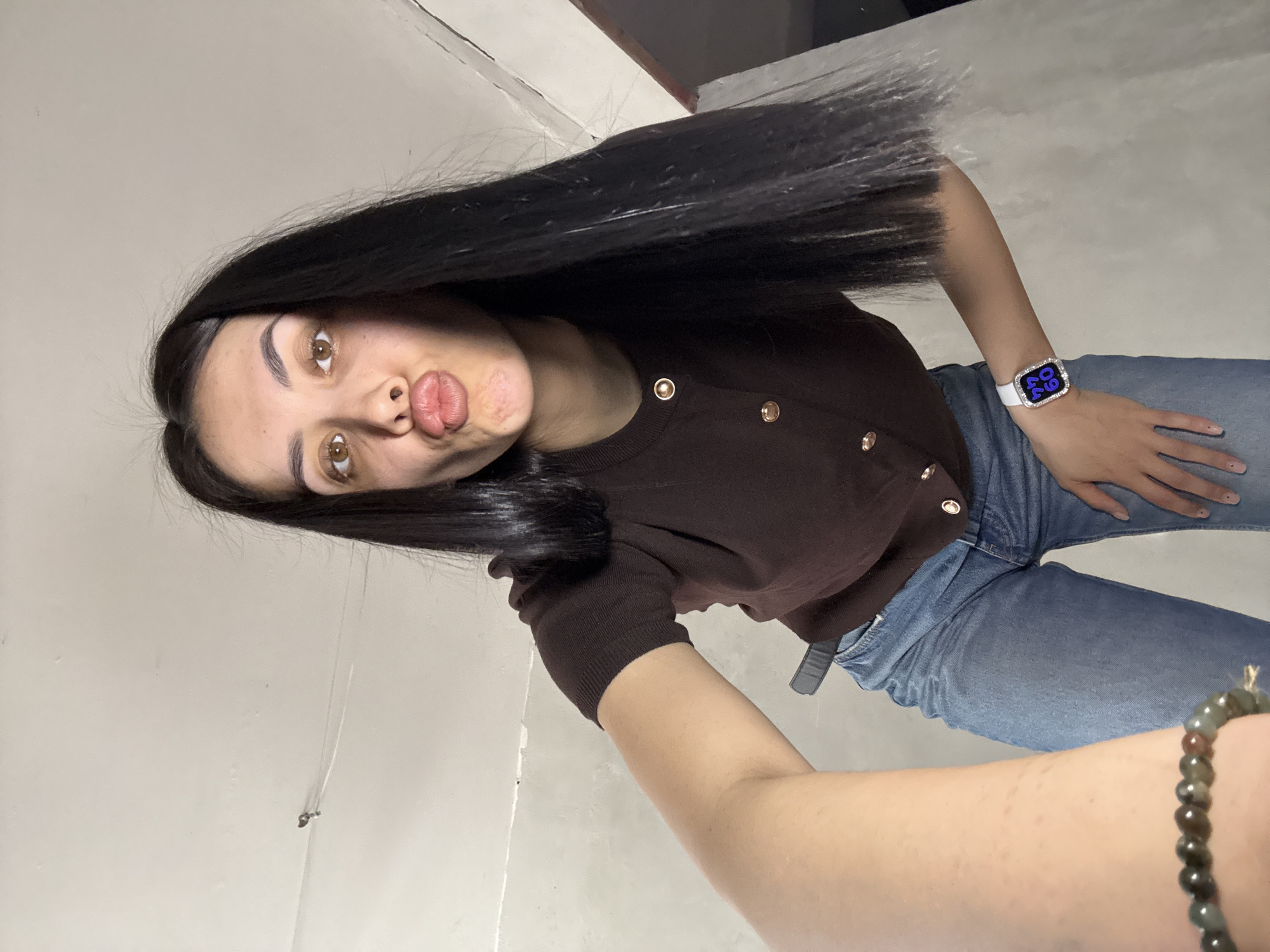 A young woman is taking a selfie. She is wearing a brown button-down shirt, blue jeans, and a white smart watch with a jeweled bezel on her left wrist. She is also wearing a beaded bracelet on her right wrist. Her hair is long, black, and straight. Her lips are pursed in a kiss. The background appears to be an indoor space with a white wall. It is not possible to determine the location in the city of Votkinsk, Russia from the given image.