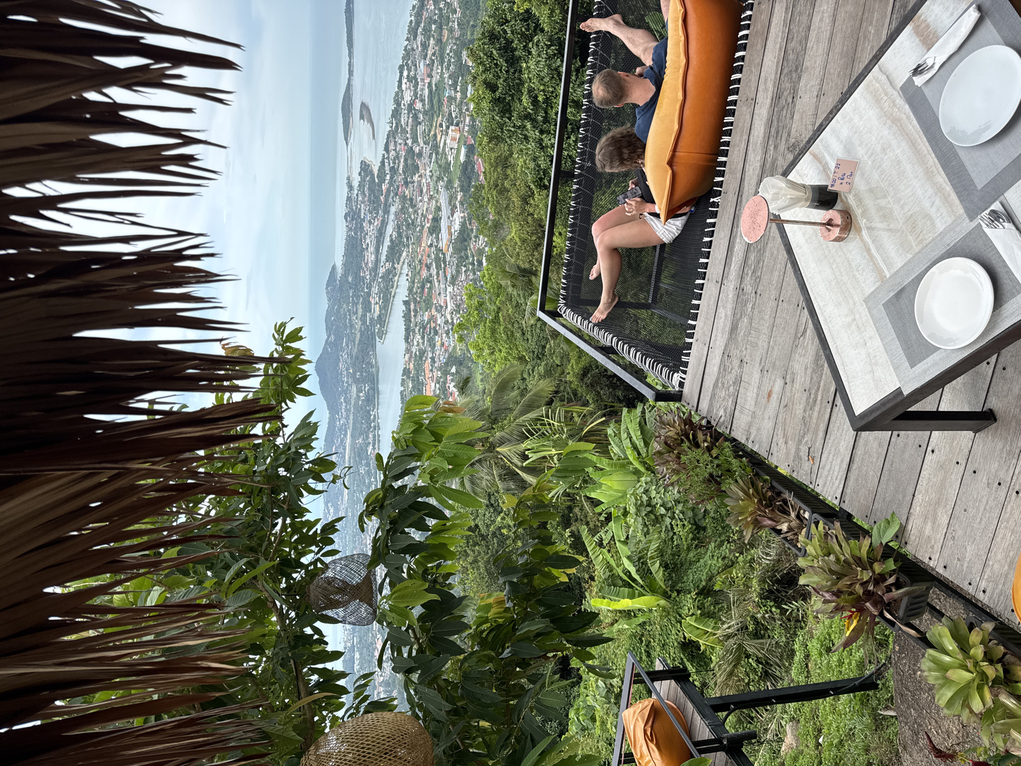 An aerial, eye-level shot captures a couple relaxing on a hammock suspended over a wooden deck, overlooking a sprawling coastal city. The man, with his back to the camera, wears a dark blue t-shirt and shorts, while the woman, facing left, sports a white top and shorts. Both are seated, with the woman holding a phone. The hammock, made of black netting, has a large orange cushion at one end. The wooden deck, weathered and showing visible grain, is in the foreground, with a glass-topped table set for two. The table has white plates, cutlery, and a small copper lamp. Behind the deck, lush green vegetation and palm trees dominate the landscape, leading to a panoramic view of the city nestled between hills and the ocean. The sky is overcast with a light grey hue.
