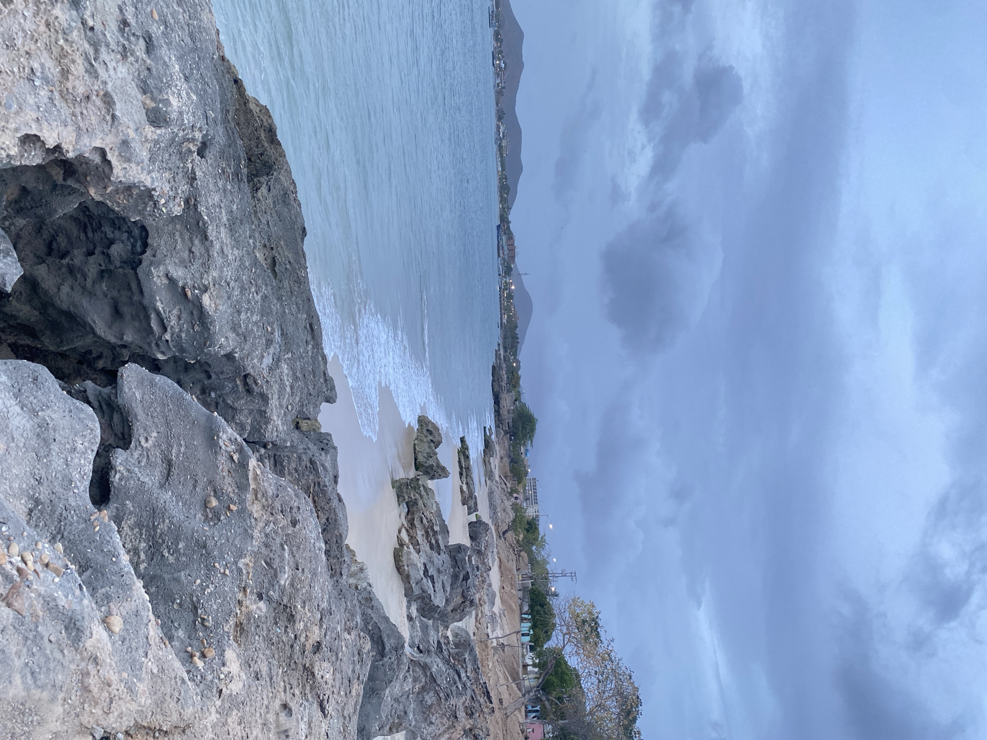 The image shows a coastal view from a rocky vantage point near Juan Griego, Venezuela. The foreground is dominated by large, grey rocks. A sandy beach stretches along the coastline, bordered by the ocean, with waves visible. Inland, there are sparse trees and some small buildings. Further in the distance, the terrain rises into mountains under a cloudy sky. The light suggests either dawn or dusk.