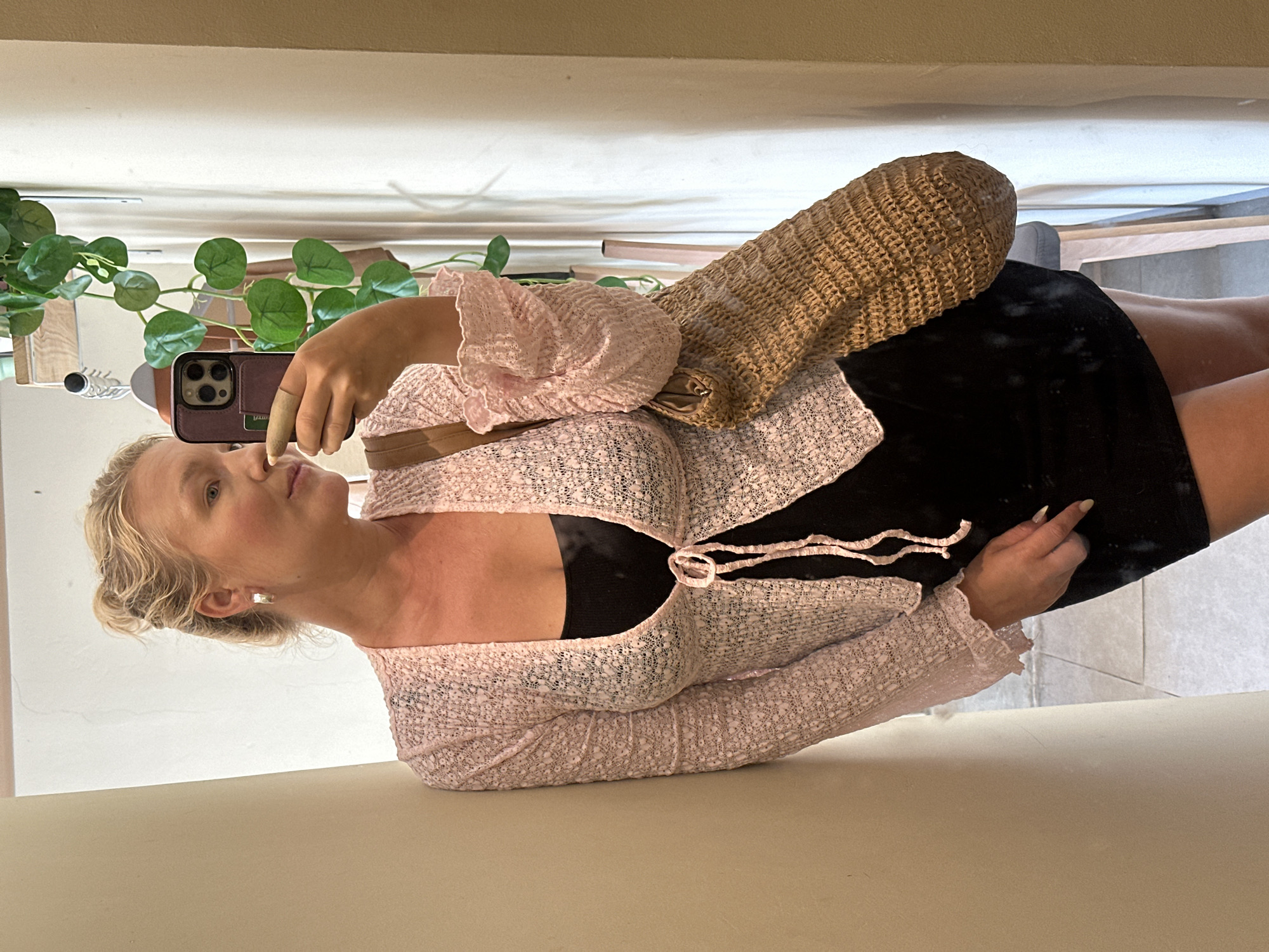 An adult Caucasian female, with blonde hair styled in a messy bun, is taking a mirror selfie. She is wearing a light pink, textured, open-front cardigan with ruched sleeves, tied at the waist with a matching pink ribbon, over a black sleeveless dress. She is accessorized with silver hoop earrings and is carrying a woven straw bag over her left shoulder. The woman is leaning against a cream-colored pillar, with her right hand holding a smartphone to take the photo. In the background, a plant with green leaves hangs from above, and a white curtain is visible behind it. The floor appears to be tiled. The setting suggests an indoor boutique or shop. The overall scene is casual and stylish. The specific location is identified as North Kuta, Indonesia.