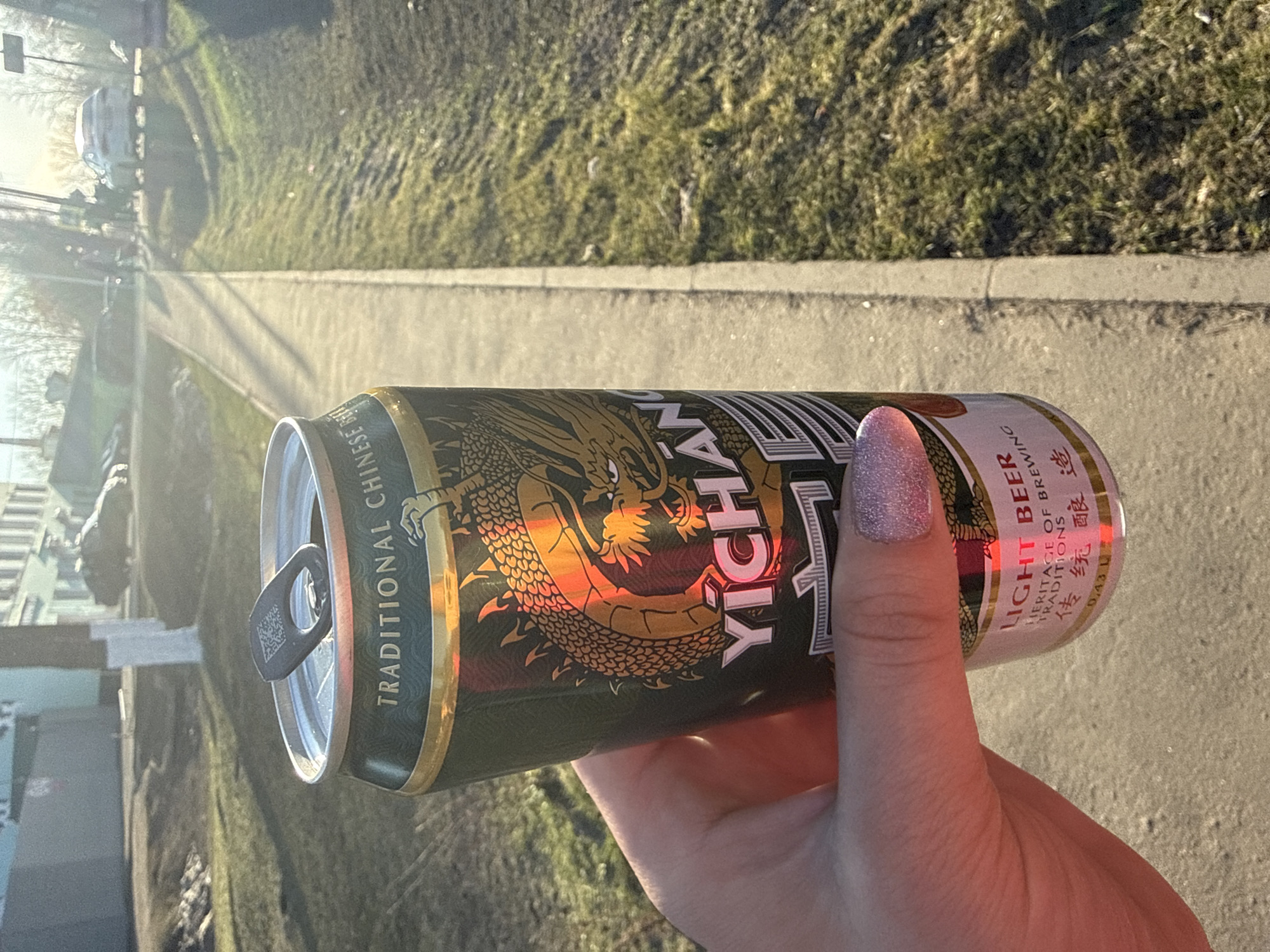 The image shows a hand holding an open can of Yichang Light Beer. The can has a design featuring a dragon and text in both English and Chinese. The background appears to be an outdoor setting, possibly in the city of Dmitrov, Russia, with grass, a sidewalk, and buildings visible.