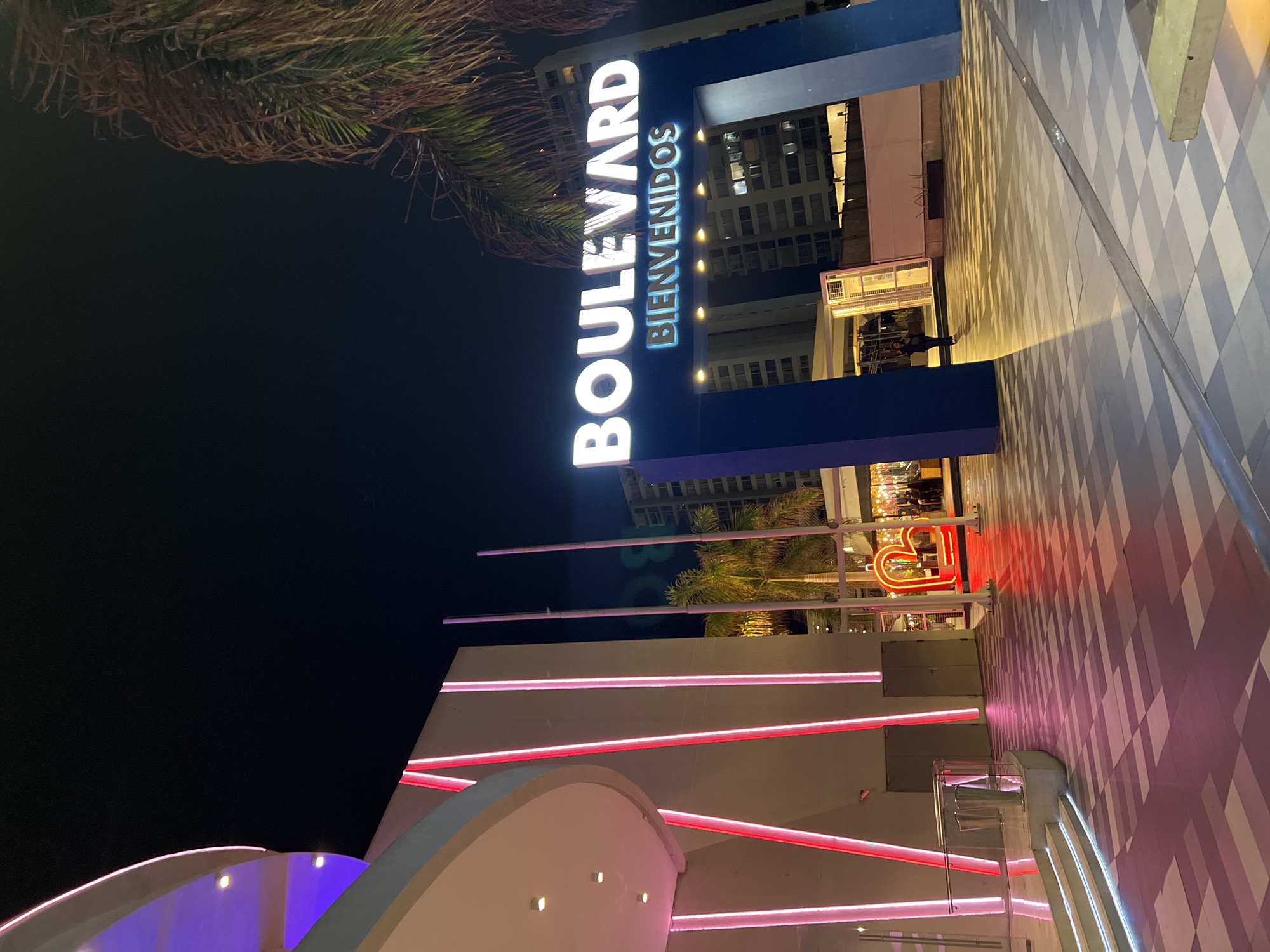 The image shows the entrance to the Boulevard shopping center in Arica, Chile, at night. The "BOULEVARD" sign is prominently displayed in white letters, with "BIENVENIDOS" in blue underneath. The sign is part of a blue structure. The building features pink and red neon lights. There is a checkered patterned walkway adjacent to the entrance. Some palm trees are partially visible.