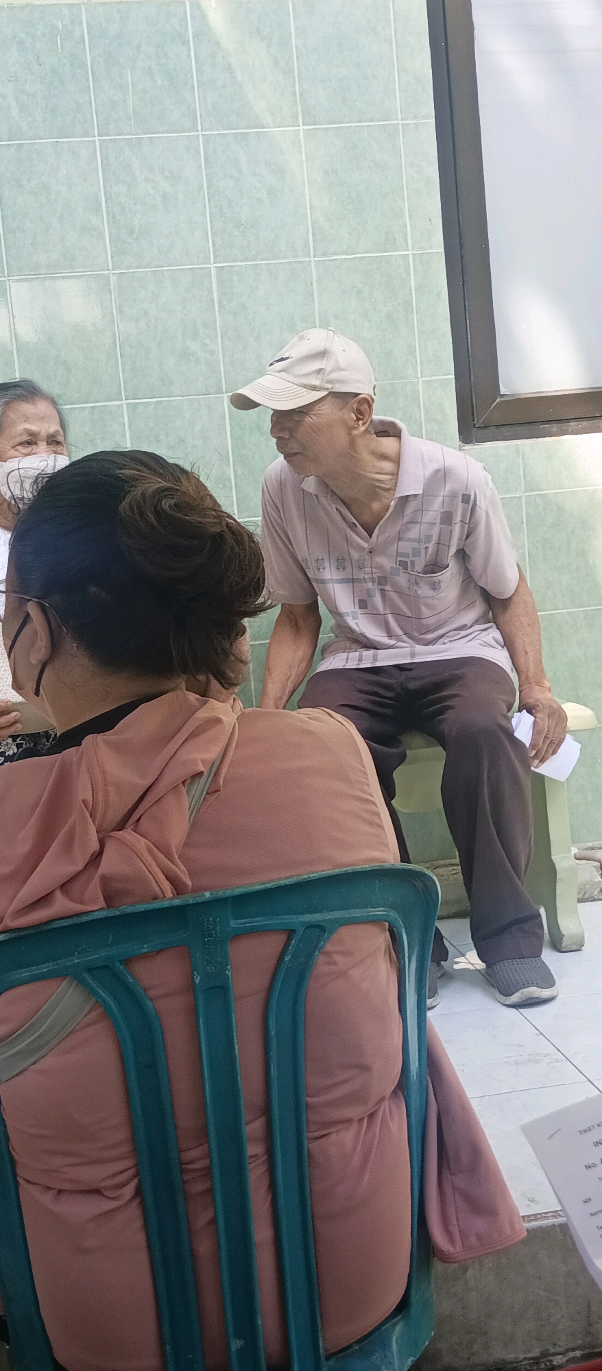 The image depicts an outdoor scene, likely in a public area, with a light green tiled wall in the background. In the foreground, a person is seated on a teal plastic chair, seen from behind, wearing a salmon-colored hooded garment. To the left, another person, partially visible and facing away, wears a white face mask and a patterned top. To the right, an elderly man wearing a light-colored baseball cap and a light-colored polo shirt with a checkered pattern is seated on a bench. He is holding a white piece of paper in his right hand. The ground is tiled. The scene appears to be a casual gathering or waiting area. The image does not contain any specific visual cues to pinpoint the location to the city of Malang, Indonesia.