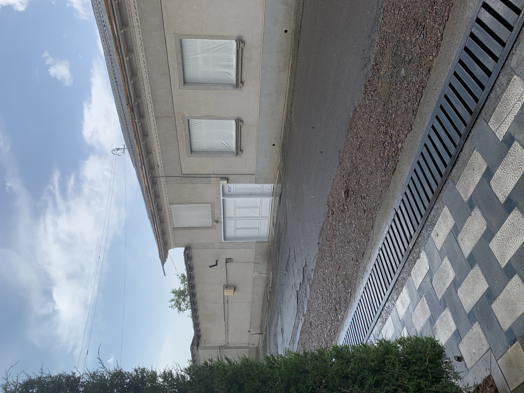 An outdoor, eye-level shot captures a street in Urtaaul, Uzbekistan, with a large building on the right and a concrete wall on the left. A green tree is on the far left. The building, constructed of grey concrete, features a dark, corrugated roof with decorative trim along the facade. Two windows on the building are covered with white sheeting. A large, white double door is visible on the right side of the building, and a smaller, single door is to its left. The street in front of the building is paved with asphalt and shows signs of wear, with patches and puddles, suggesting recent rain. A drainage grate runs along the edge of the street, adjacent to a patterned walkway made of grey and white tiles. The background shows a cloudy, pale blue sky with some faint electrical wires strung across it.