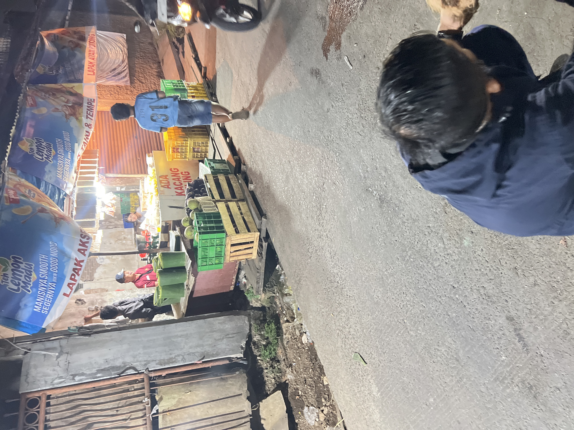 The image depicts a street scene at night in Citeureup, Indonesia, likely a market or roadside stall area. In the foreground, a person wearing a dark blue jacket is crouched down, looking towards the right. A small portion of a brown dog is visible near this person. In the midground, a young person in a blue t-shirt with the number 31 on the back is walking away from the camera. Behind them is a stall with a blue tarp overhead advertising "Teh Lemon Madu" (Lemon Honey Tea). Various goods are displayed at the stall, including green crates filled with produce, possibly durians, and other items in wooden crates. Two individuals are visible behind the counter of the stall. To the right, a motorcycle is parked, its headlight illuminated. The ground is paved and shows signs of being a public thoroughfare, with some debris and a drainage area visible on the left. The lighting suggests it is nighttime.