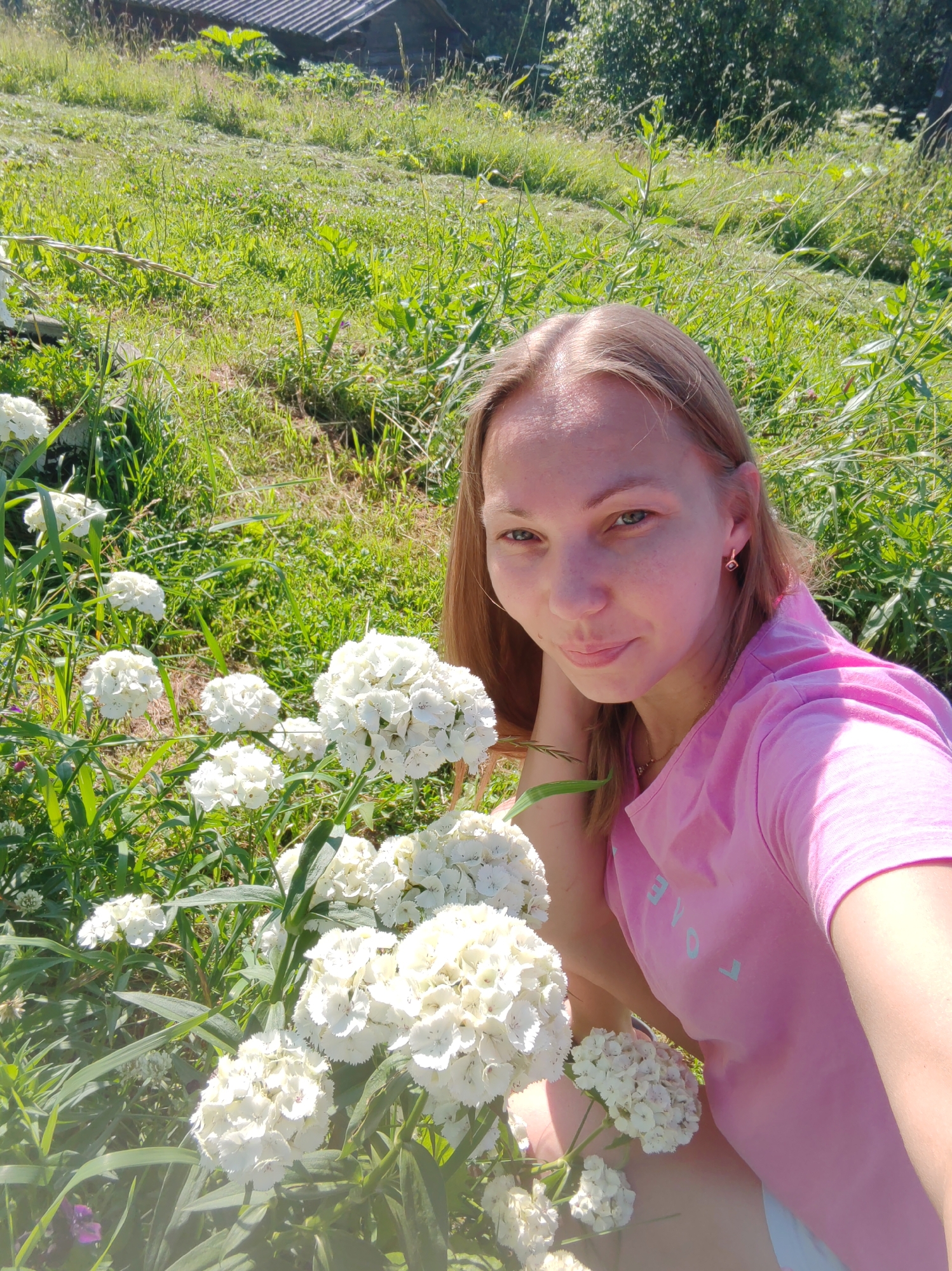 A young woman is crouching in a grassy field with white flowers in the foreground. She is wearing a pink t-shirt and has long, light brown hair. The background shows a wooden structure, possibly a house, and trees. The scene appears to be set in a rural area, potentially in the city of Chukhloma, Russia, during the daytime. The woman is looking at the camera with a slight smile.