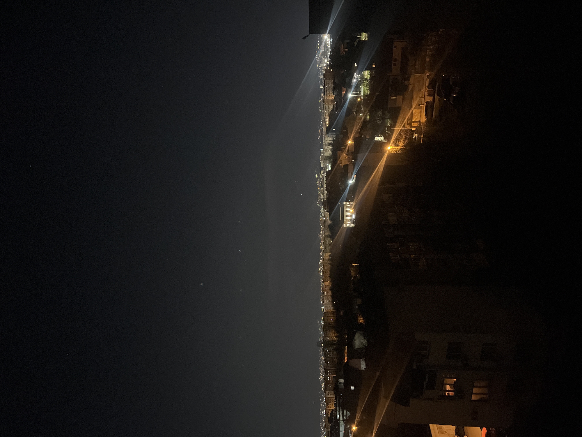 The image shows a nighttime view of Sofia, Bulgaria. The left side of the image is predominantly dark, representing the sky. A few small, faint lights are visible, possibly stars. The right side of the image shows the city, with numerous lights from buildings and streets. The lights create streaks and patterns that illuminate the urban landscape. Buildings of varying heights are visible, forming a dense urban environment. Some buildings in the foreground are more clearly visible, with illuminated windows. The overall scene is a contrast between the dark night sky and the bright lights of the city.