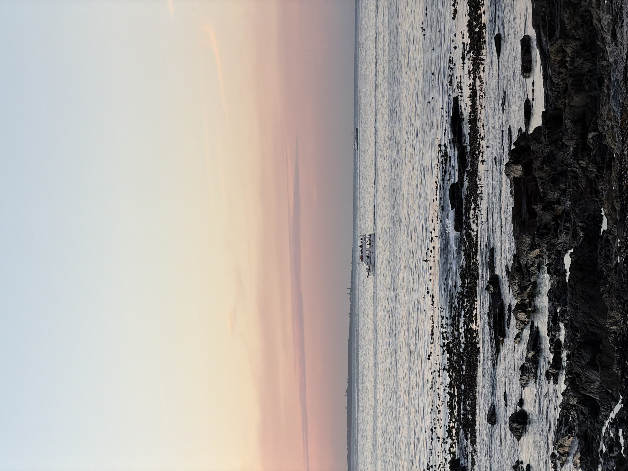 The image depicts a coastal scene in Cape Town, South Africa at either dusk or dawn. On the left side of the image is the sky, which transitions from light blue to orange and pink hues. A narrow sliver of land separates the sky from the water. In the center of the image, a boat is visible on the water, carrying people. To the right of the boat is the ocean, and on the far right are rocky formations along the shoreline.