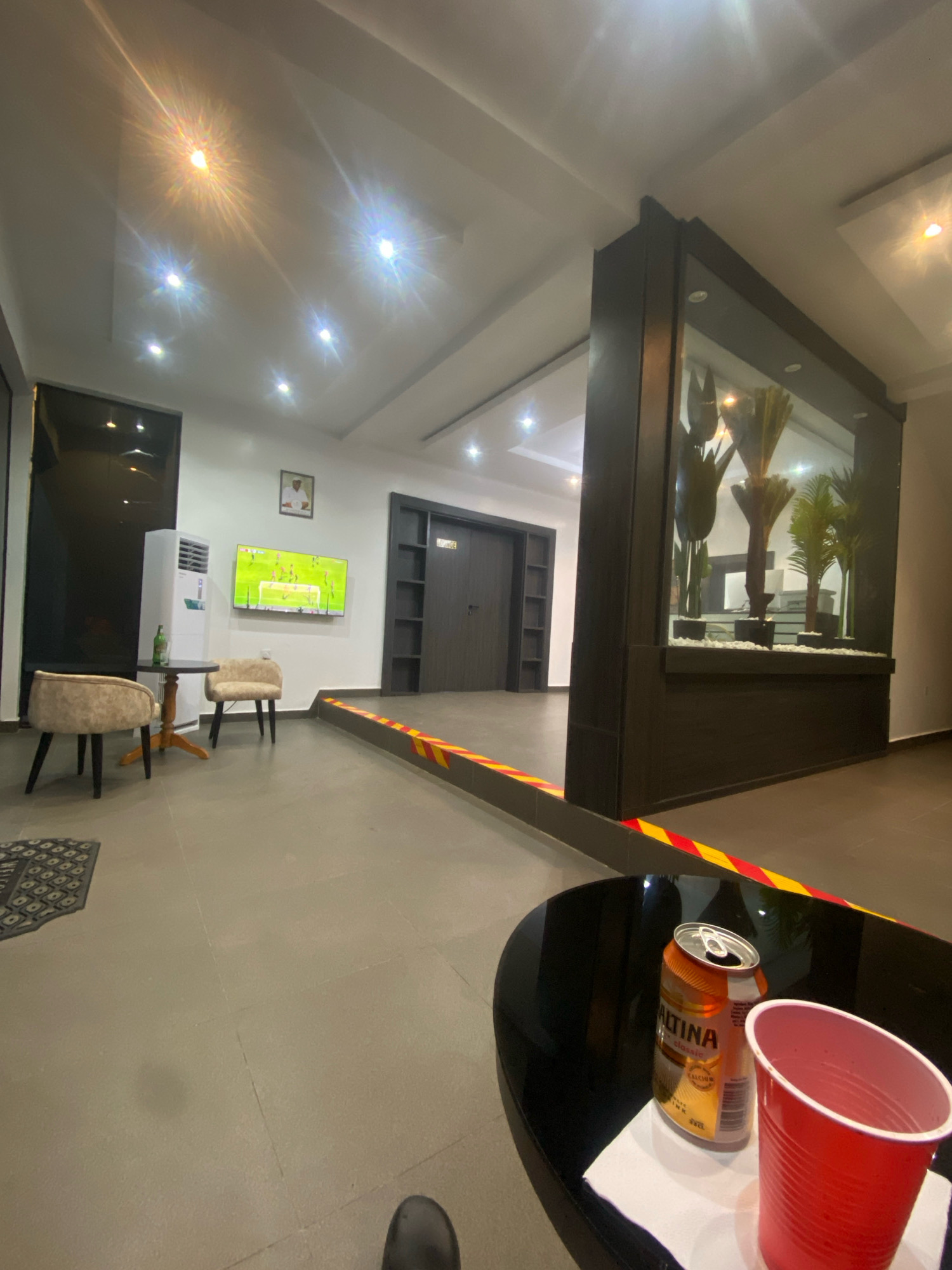 The image depicts an indoor scene, likely a lounge or common area, with a contemporary design. The space features a large television screen displaying a soccer match, two beige upholstered chairs, and a small round table with a white air conditioning unit and a bottle on it. A dark, rectangular pillar with a built-in glass enclosure containing artificial plants is a prominent feature. The floor is tiled with large, light-colored squares. Yellow and red hazard tape is visible on the floor, indicating a boundary or caution area. In the foreground, a black coffee table holds a partially opened can of "ALTINA classic" drink and a red plastic cup. The lighting appears to be from overhead recessed fixtures, some of which are creating lens flares. The location is identified as Barden Yana, Nigeria.