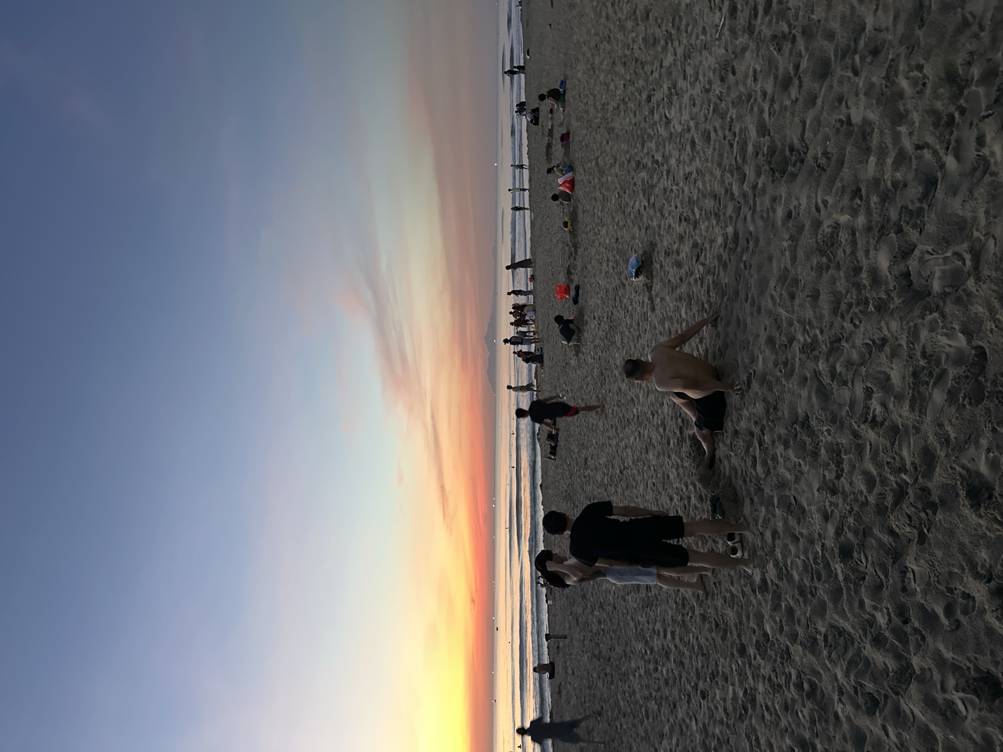 The image depicts a wide shot of a beach at sunset. The sky is painted with vibrant hues of orange, pink, and yellow, transitioning into a soft blue. The sun is low on the horizon, casting a warm glow over the scene. The beach itself is sandy, with numerous footprints visible, indicating activity.
Several people are scattered across the beach, engaged in various activities. Some are walking, while others are standing or sitting. In the foreground, a group of three people are the most prominent, with two individuals embracing. Further back, a man is captured mid-stride, appearing to be running or exercising. Other figures are visible in the distance, closer to the water's edge, some of whom seem to be playing or wading in the shallow waves. The ocean stretches out to the horizon, with gentle waves rolling onto the shore. The overall atmosphere appears relaxed and serene, typical of a beach at dusk. There is no visible text in the image.