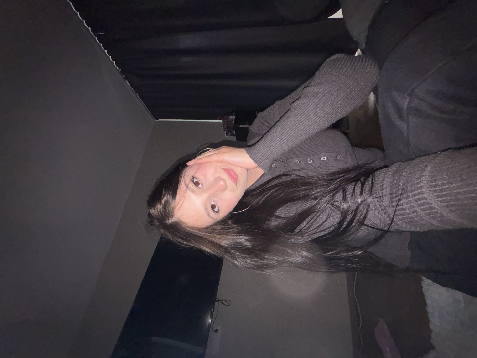 An indoor, tilted portrait features a young woman with long, dark hair, likely taken in Chaikovsky, Russia. She is positioned diagonally in the frame, looking directly at the camera with a neutral to slightly smiling expression. Her right hand is raised, with fingers gently touching her face near her eye. She is wearing a dark grey, ribbed long-sleeved top with small buttons running down the front. Gold hoop earrings are visible.
The setting appears to be a dimly lit room, possibly a bedroom or living area. In the background to her left, a dark, switched-off television screen is visible, with some wires below it. The walls are a dark grey, and behind her to the right, there are dark fabrics, possibly curtains or upholstery of a bed/couch, which are also dark grey or black. The ceiling is dark and textured. The overall lighting suggests it is either evening or night, relying on minimal ambient light. No discernible text is visible in the image.