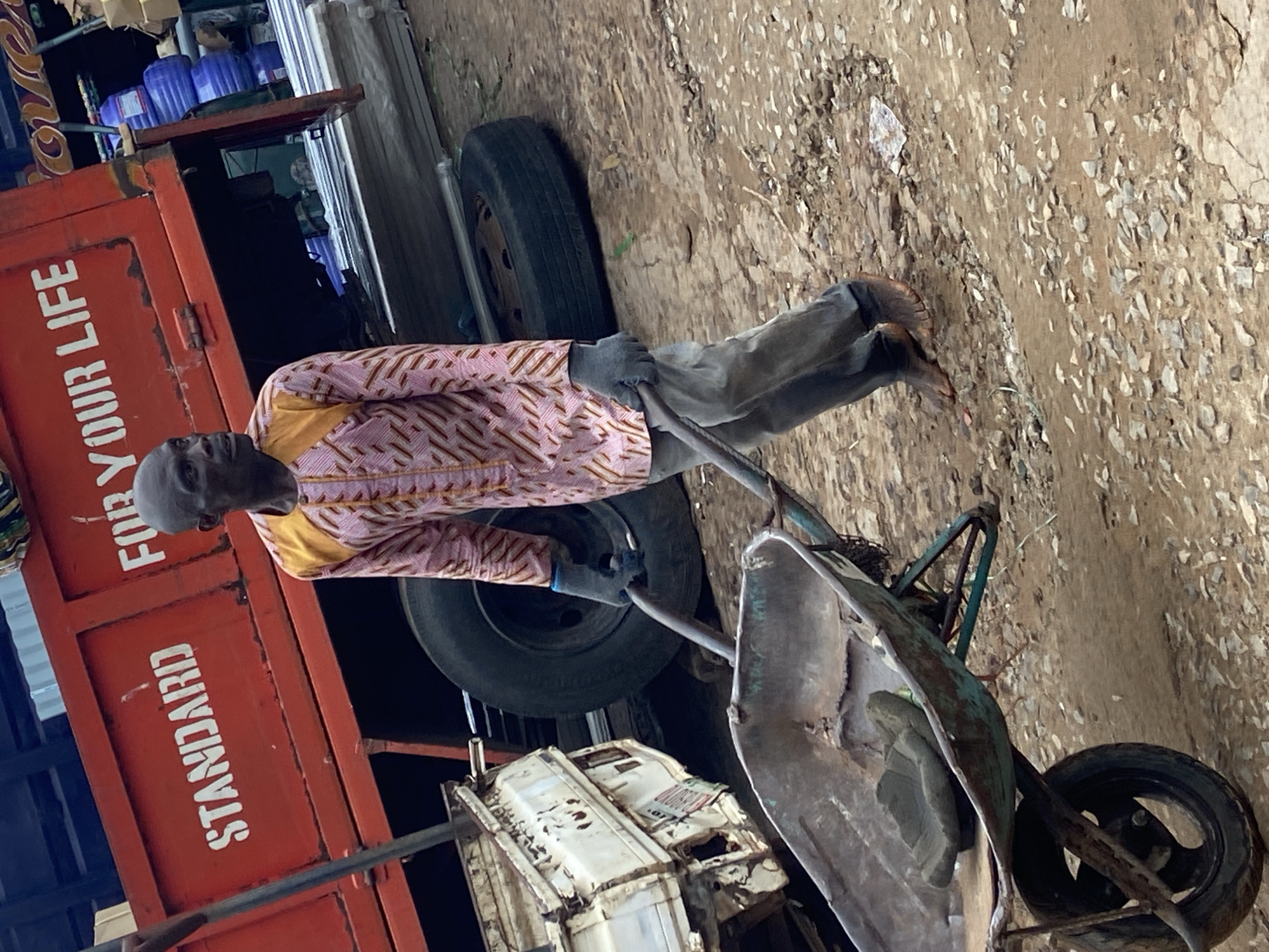 In Ogbunike, Nigeria, a man is pushing a wheelbarrow. He is wearing a pink and gold patterned garment and gray trousers. His arms are gloved, and he is leaning forward, his face turned upwards and slightly to the left. The wheelbarrow appears to be carrying a load covered by a tarp. Behind the man, a red truck with the words "FOR YOUR LIFE" and "STANDARD" painted in white on its side is visible. The truck is loaded with blue water bottles. The scene is outdoors, with a dirt and gravel ground. The lighting suggests it is daytime.