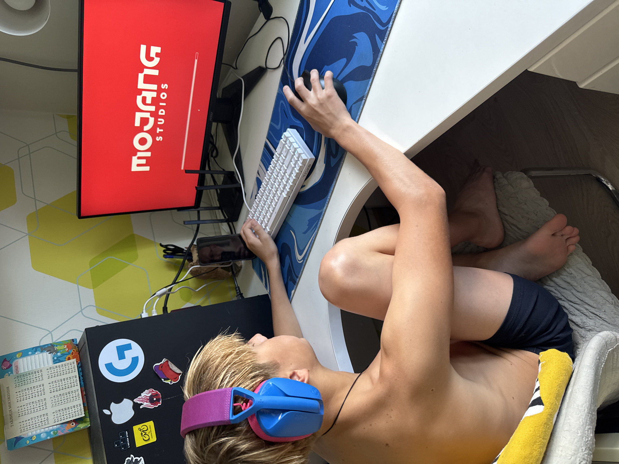 The image depicts a young person, likely a boy, seated at a desk in a room. He is wearing blue and pink headphones and appears to be playing a video game, as indicated by his focus on the computer monitor. The monitor displays the "Mojang Studios" logo on a red background. On the desk, there is a white keyboard, a black computer mouse, and a blue and white mousepad with a swirling design. To the left of the monitor, there is a black laptop with various stickers, including an Apple logo and a Logitech "G" logo. Above the laptop, a colorful multiplication table chart is visible. The room has patterned wallpaper with yellow and green geometric shapes. The person is shirtless and wearing dark blue shorts. The overall setting suggests a typical home environment, possibly a bedroom or a dedicated gaming space. The image does not provide any specific information or visual cues that would allow for its location to be determined as Zaporizhzhia, Ukraine, or any other specific city or region.